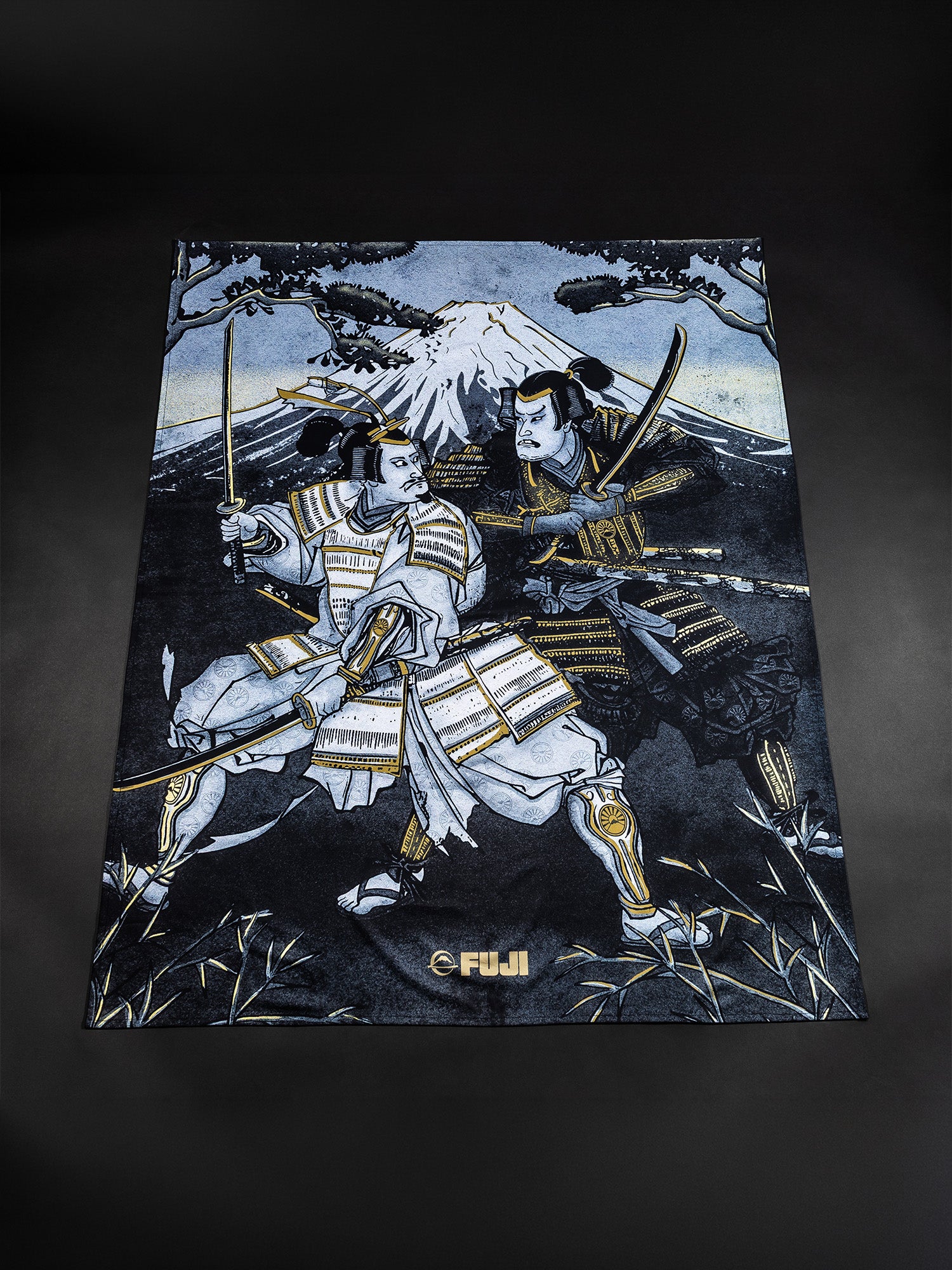 A towel from the Heritage Keishō Edition Bundle featuring an illustration of two armored samurai wielding swords, standing before Mount Fuji with tree branches overhead. The word FUJI appears at the bottom of the mostly blue, white, and gold design.