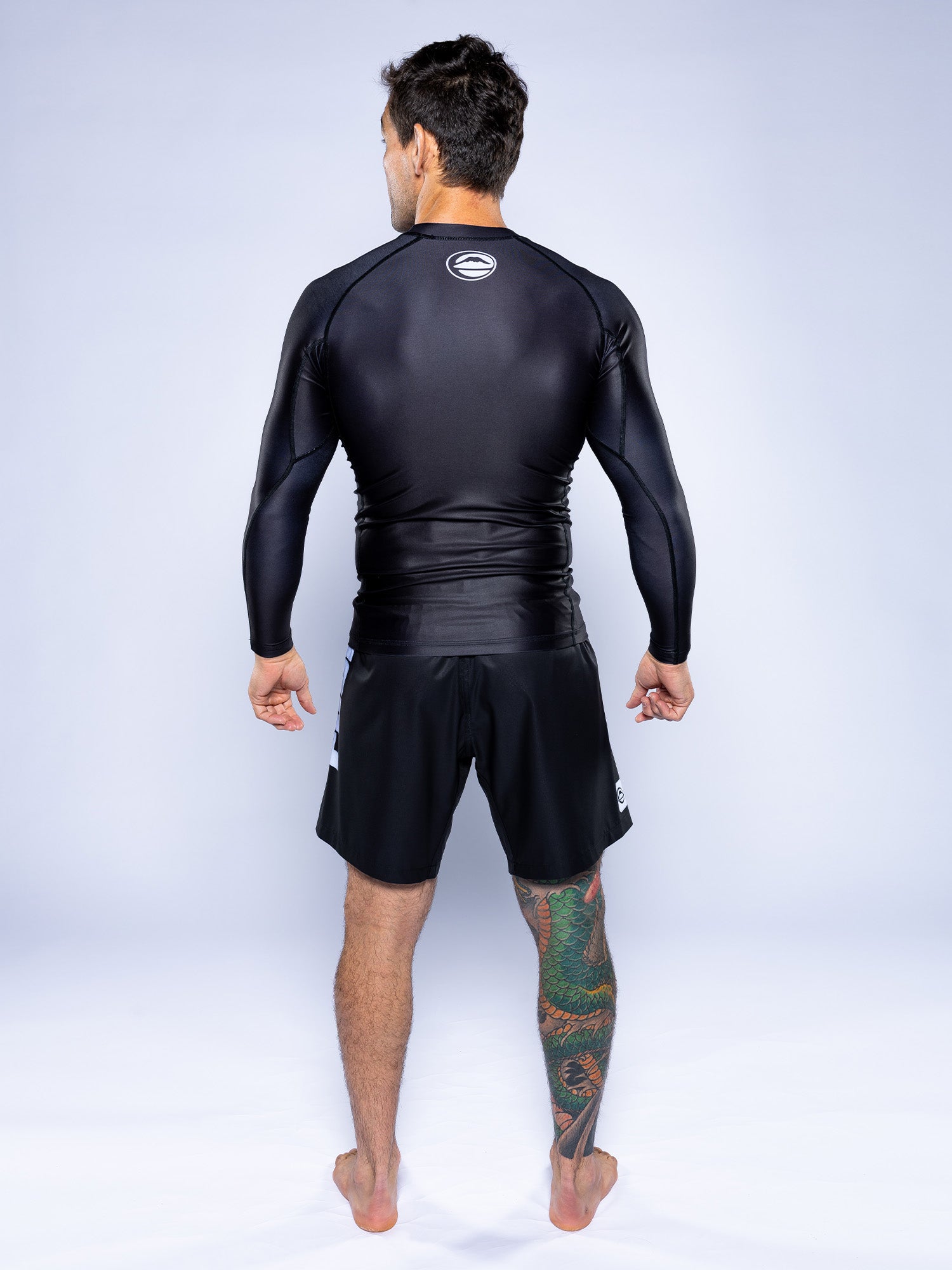 A man stands barefoot facing away, wearing the Baseline 2.0 Long Sleeve Rashguard Black and black shorts, with a large colorful tattoo on his left leg against a plain light background.