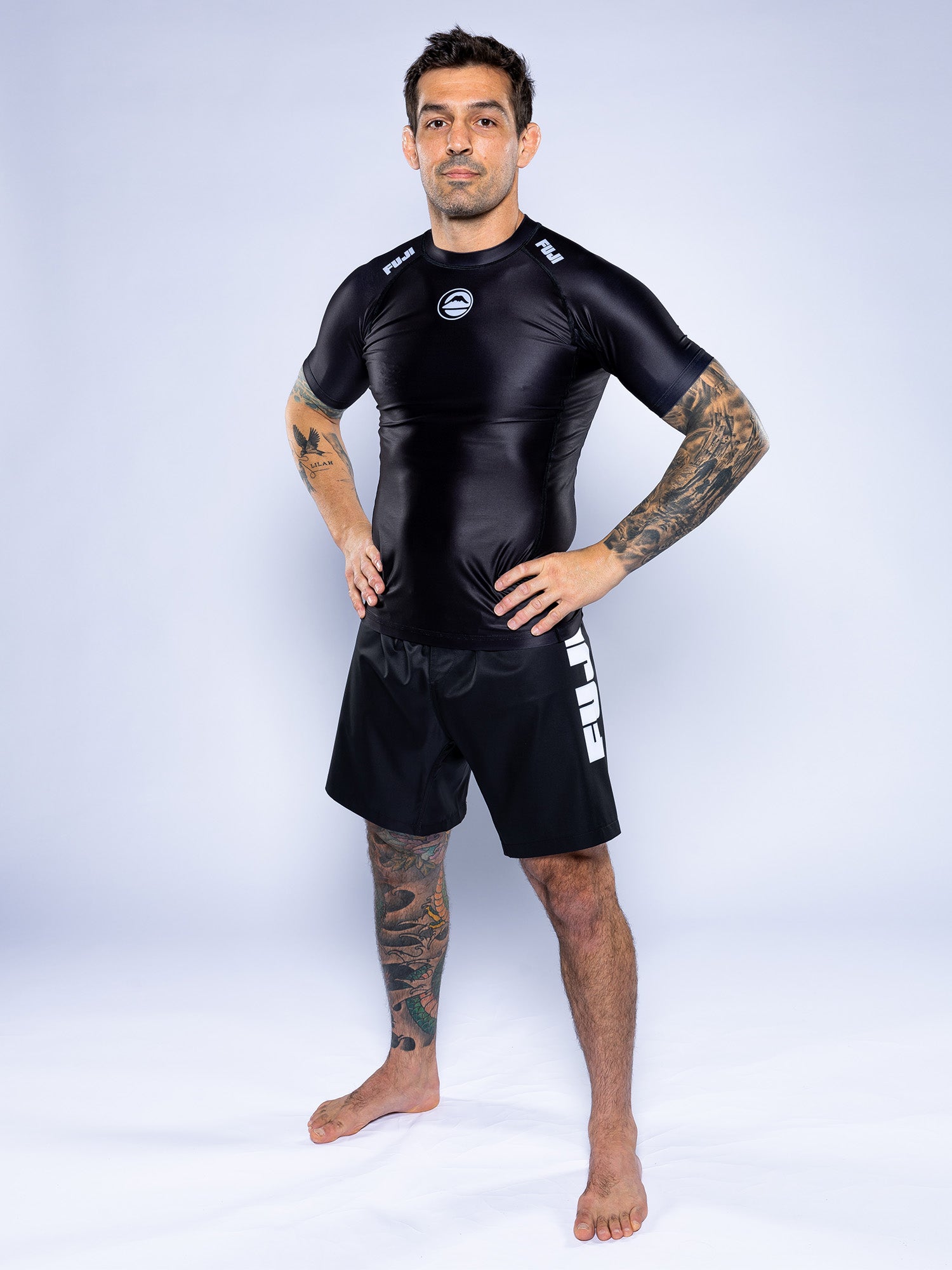 A tattooed man stands barefoot on a white surface, confidently wearing the Baseline 2.0 Short Sleeve Rashguard Black and black shorts with “FULL” on the side, his hands resting on his hips.