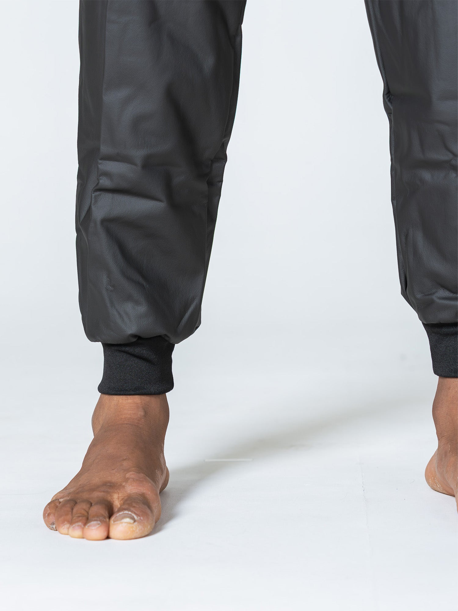 A close-up of bare feet on a light surface, wearing the Grappling Sauna Suit Black with shiny, cuffed black pants. Lower legs and ankles are visible.