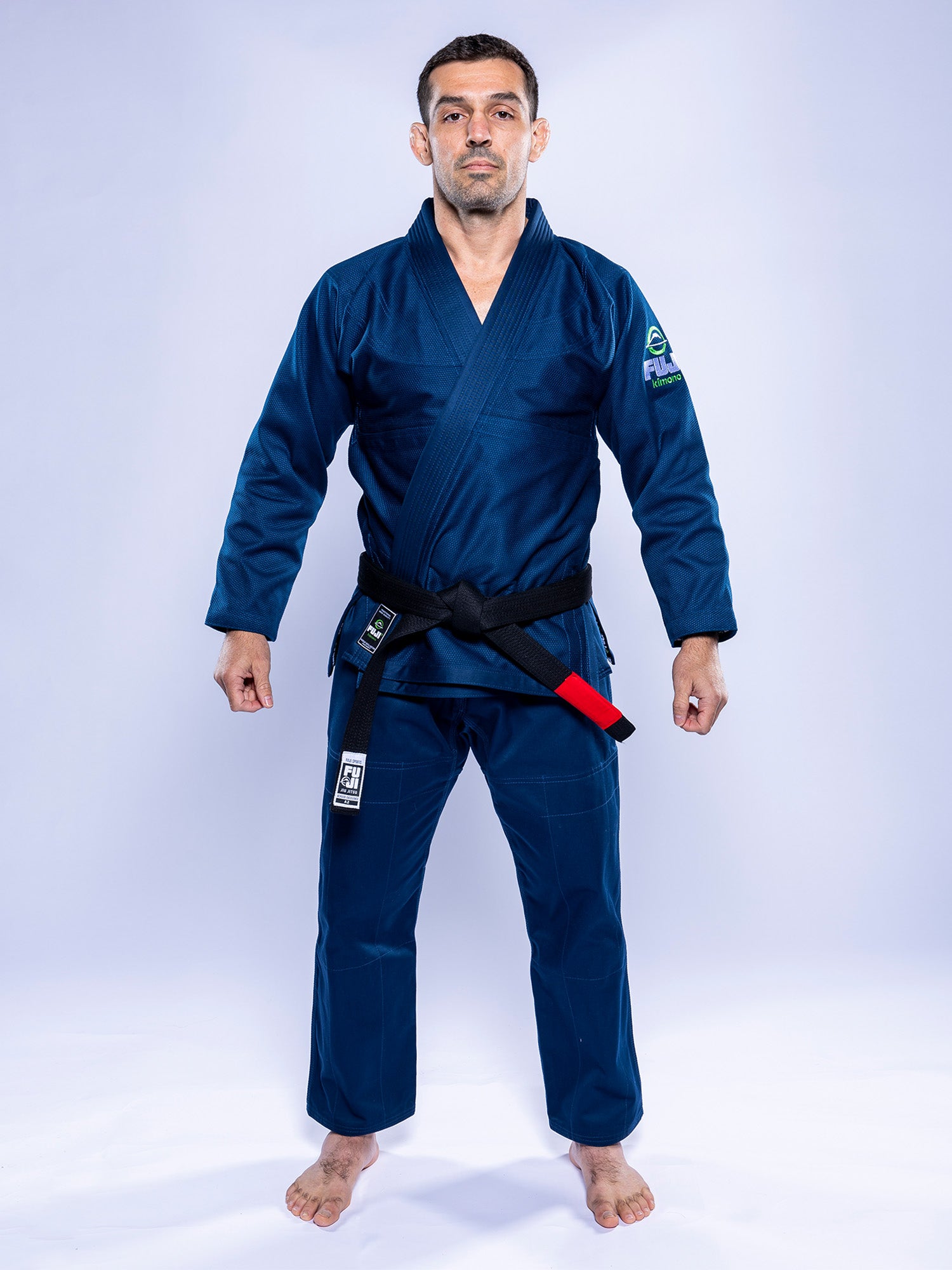 A man stands barefoot against a plain light background, wearing the Lightweight BJJ Gi Navy with a black belt. He faces forward.
