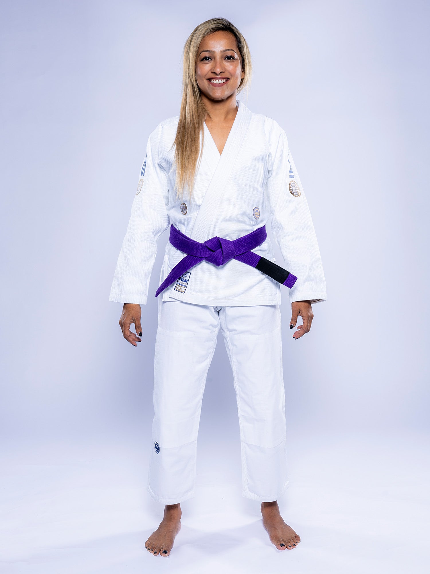 A woman stands barefoot, smiling, wearing the Womens SEKAI 3.0 BJJ Gi White Navy with a purple belt against a plain light background.