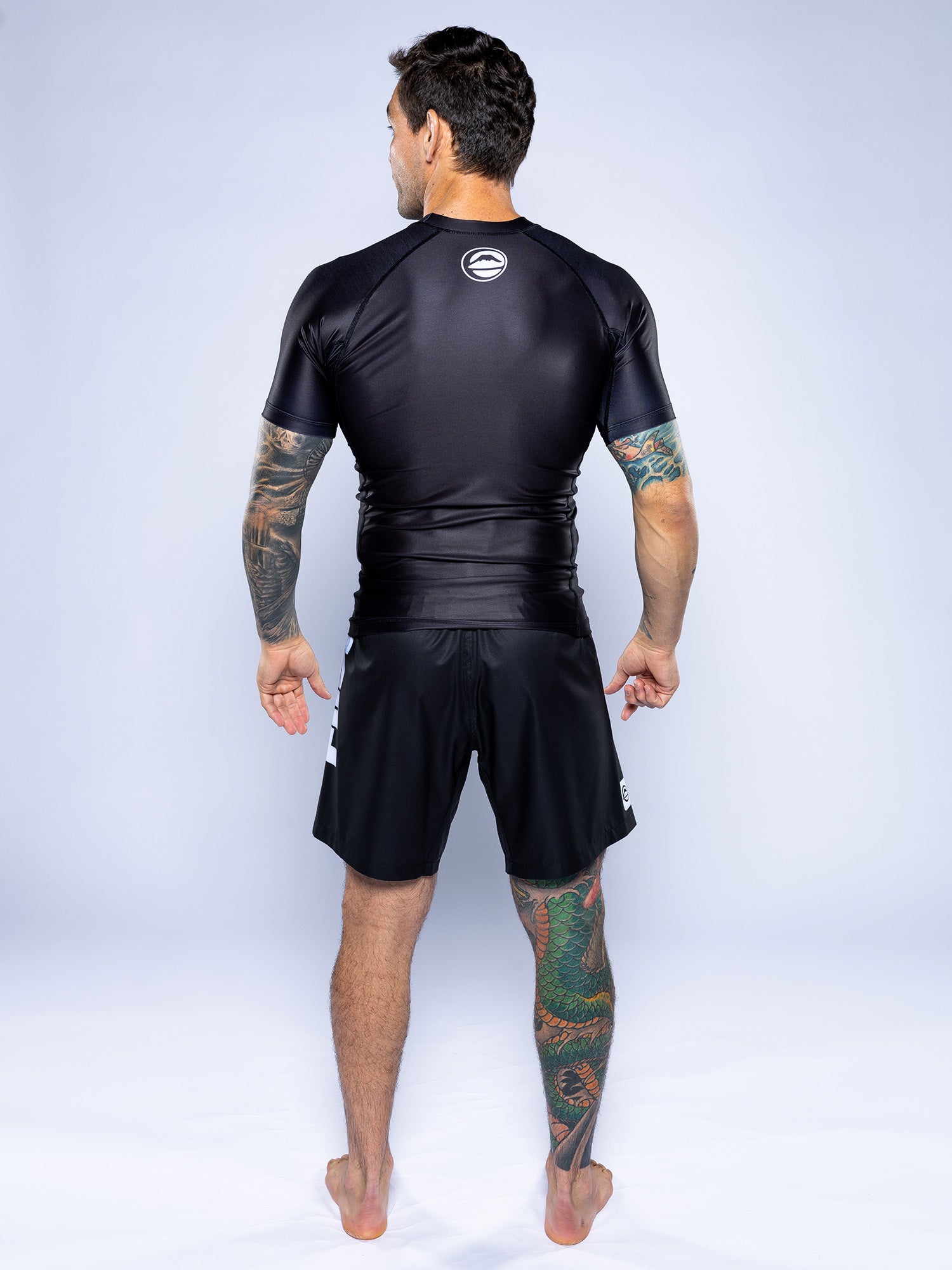 A tattooed man stands barefoot, facing away in a Baseline 2.0 Short Sleeve Rashguard Black and black shorts with white accents. He has tattoos on both arms and his left leg. The background is plain and light-colored.
