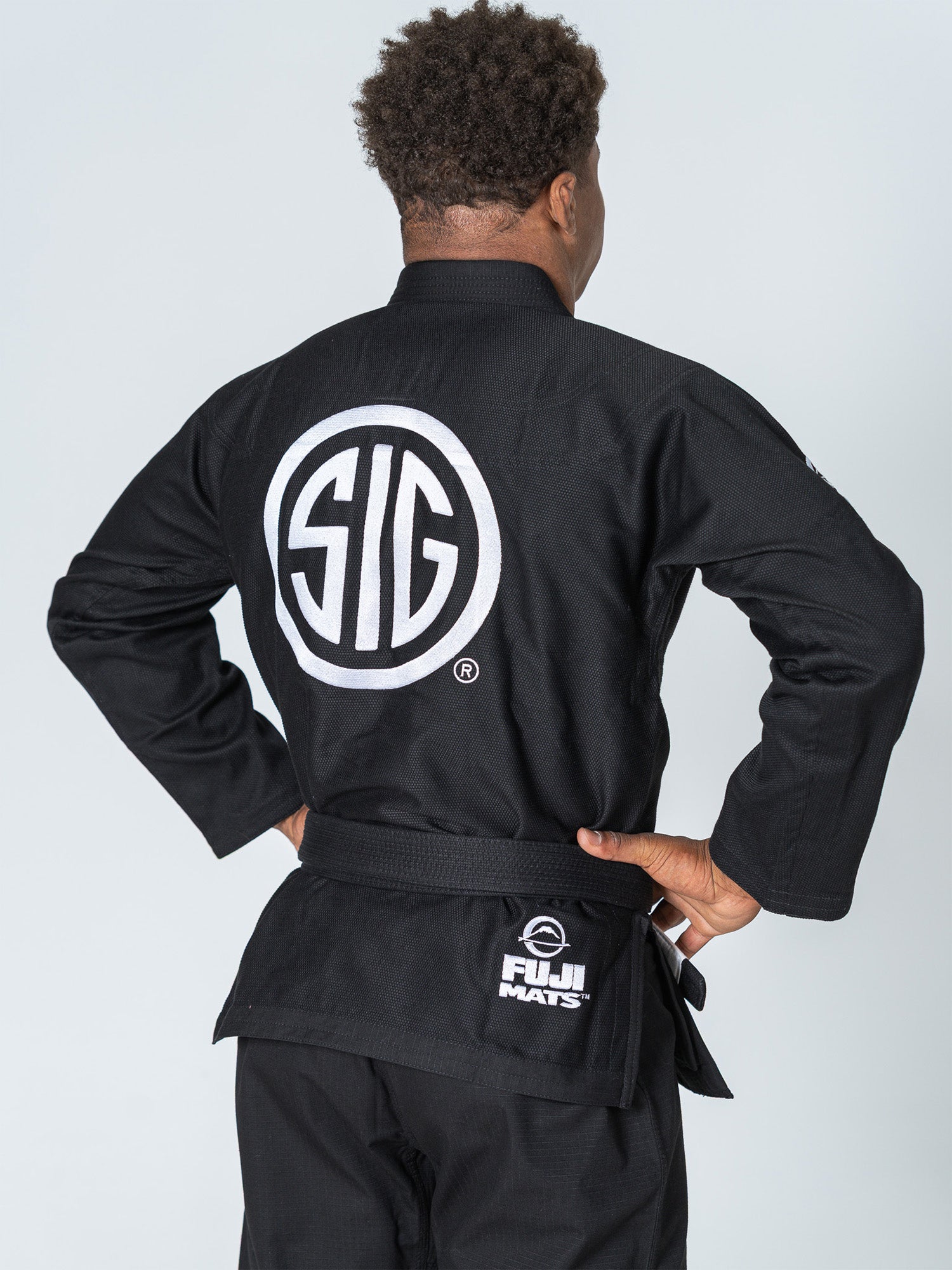 A person wearing the SIG SAUER Suparaito Gi Black with a large white “SIG” logo on the back and a “FUJI MATS” label on the belt stands facing away, hands on hips.