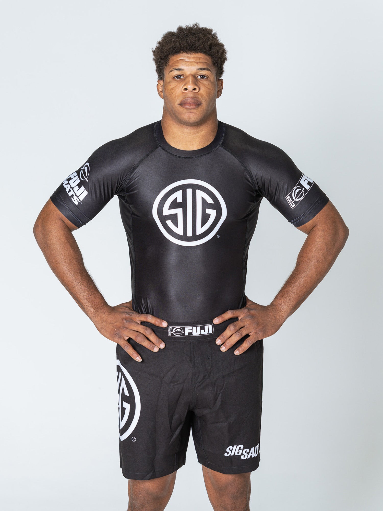A man stands against a plain background wearing the Sig Sauer Short Sleeve Rashguard in black and matching shorts with white SIG logos, hands on hips, looking directly at the camera.