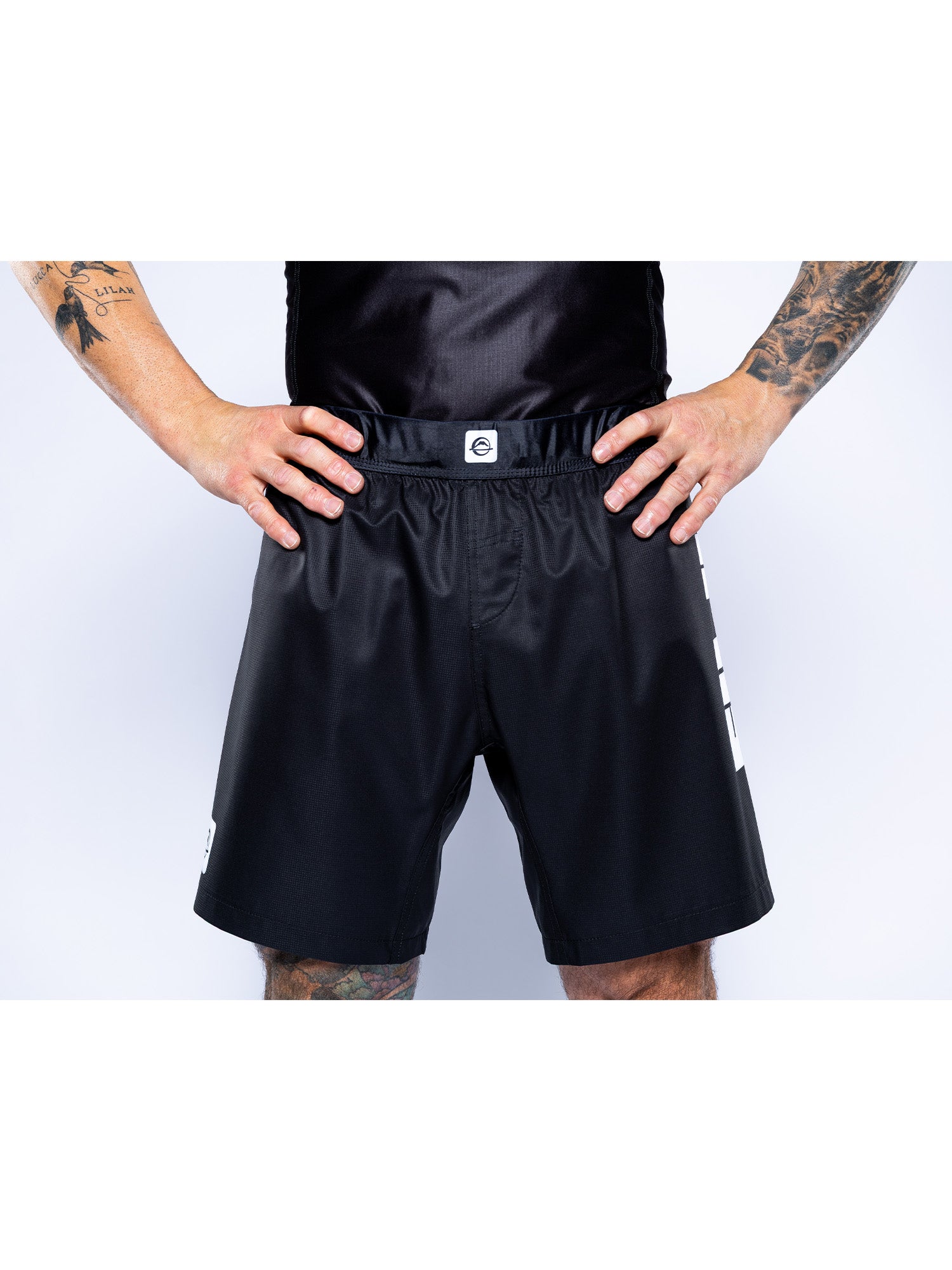 A person wearing Competition Ranked Shorts Black and a black top stands with hands on hips, showing tattooed arms and legs against a plain white background.