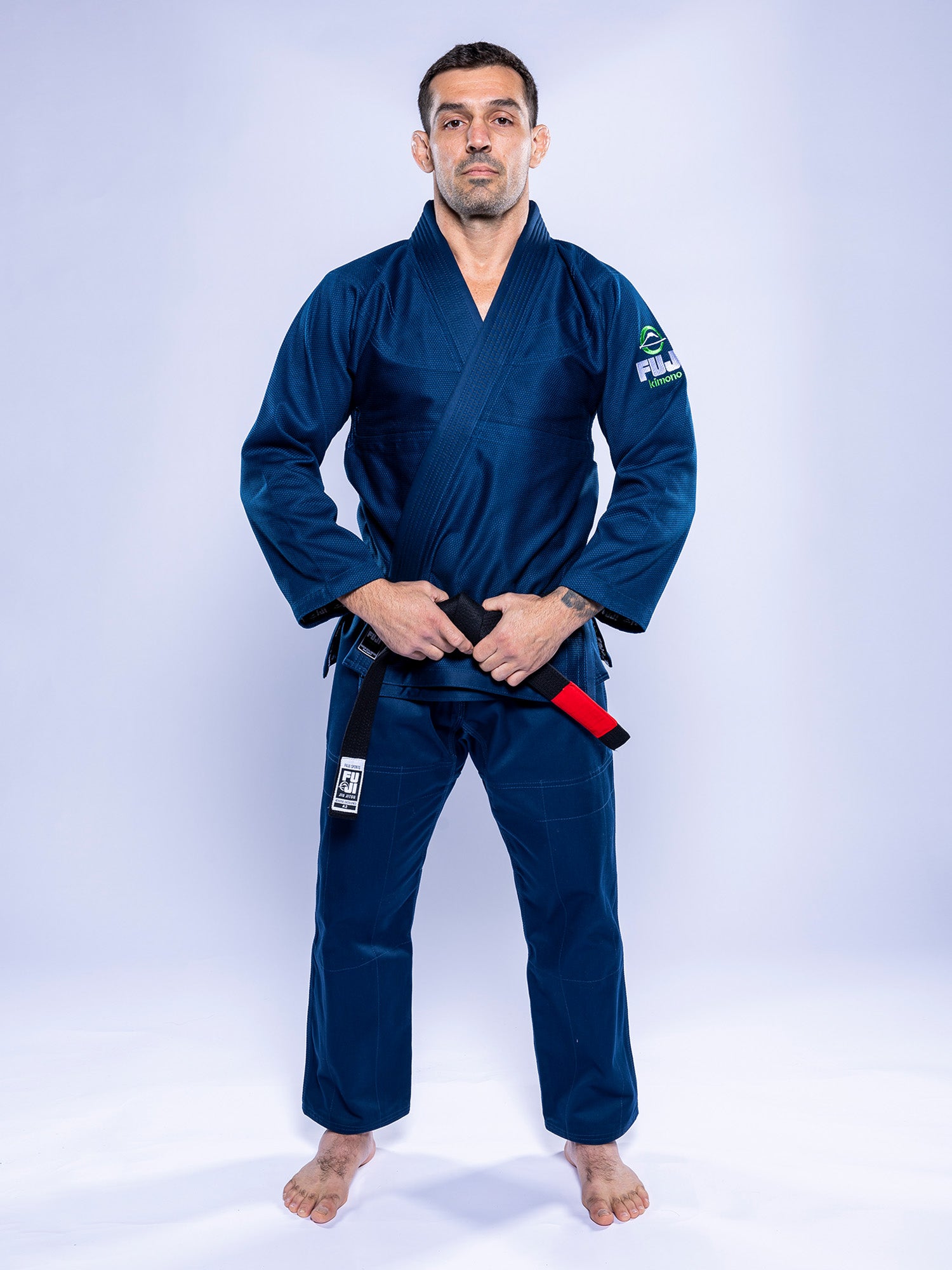A man in the Lightweight BJJ Gi Navy stands barefoot against a plain light background, facing forward with a serious expression and holding a black belt.