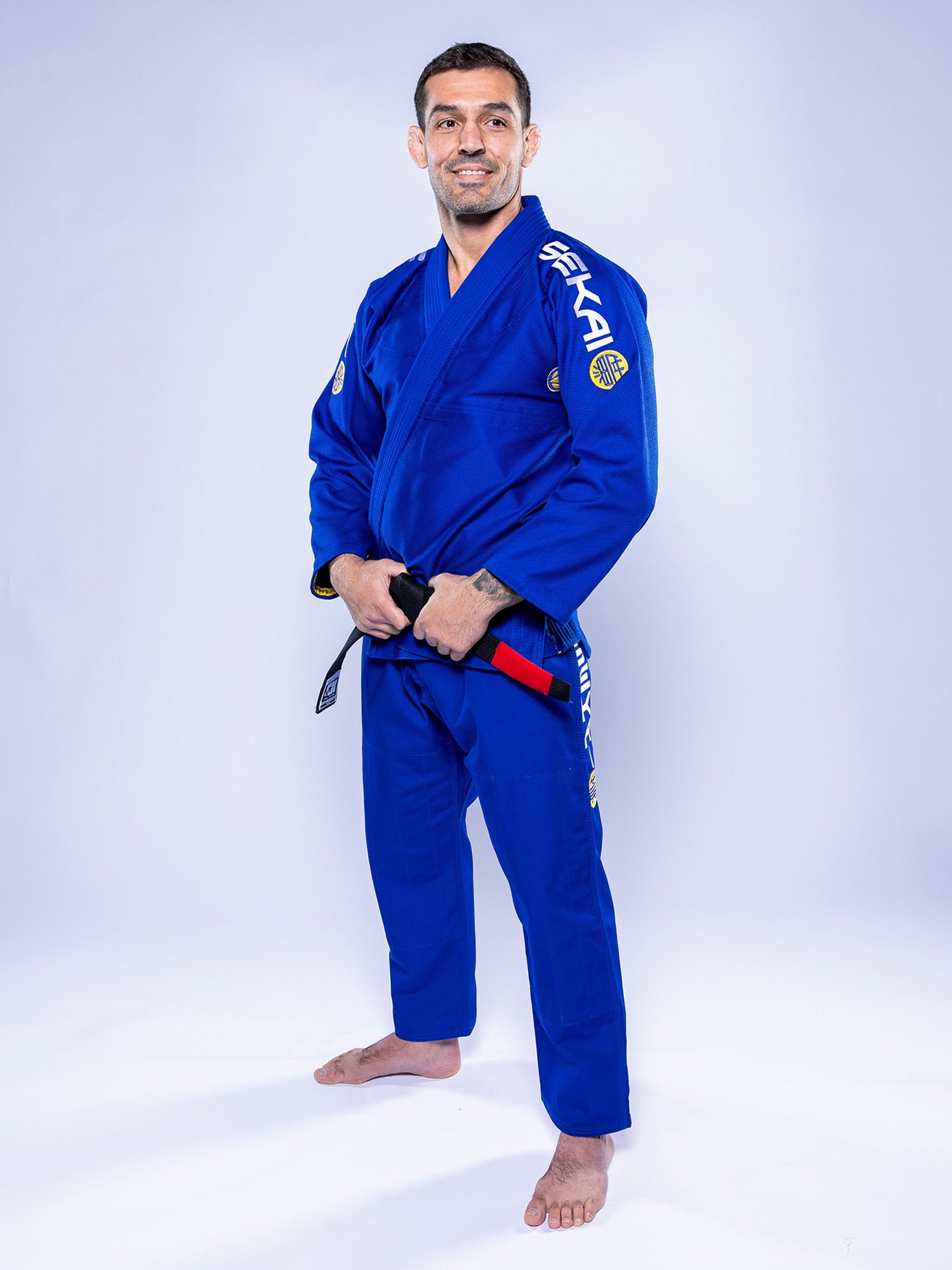 A man wearing the SEKAI 3.0 BJJ Gi Blue Yellow with a black belt stands barefoot on a white background, smiling and facing the camera.