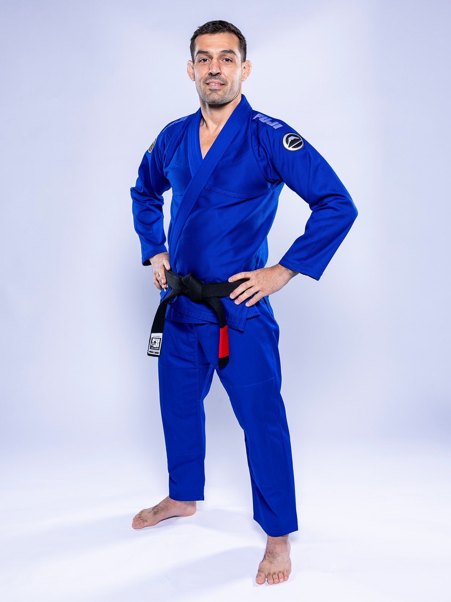 A person wears the Baseline 2.0 BJJ Gi Blue White with a black belt, standing barefoot on a white background, hands on hips, smiling at the camera.
