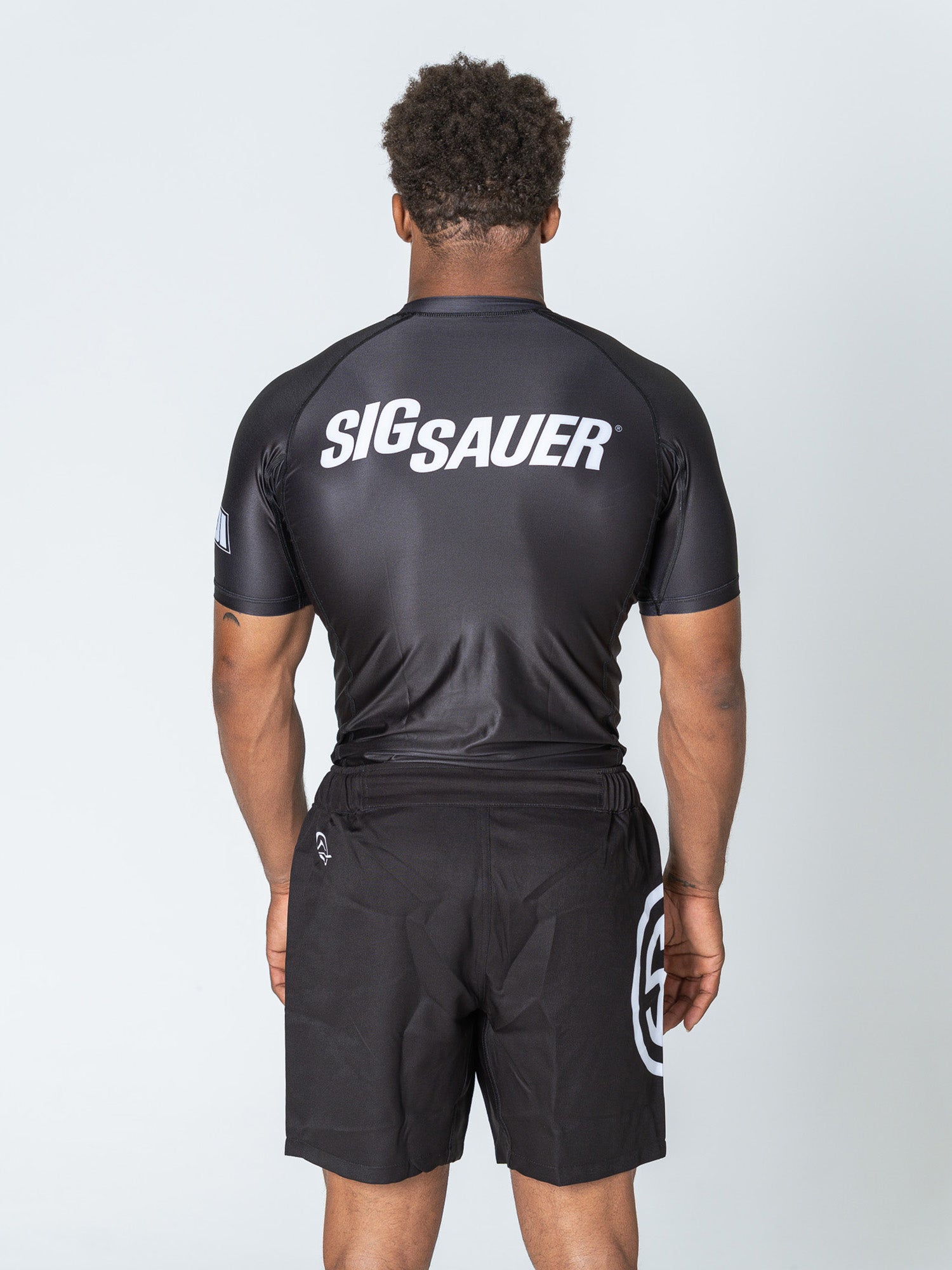 A man faces away wearing the Sig Sauer Short Sleeve Rashguard in black, featuring “SIGSAUER” in white across the back and paired with black athletic shorts with a white circular logo on the left leg. The background is plain and light-colored.