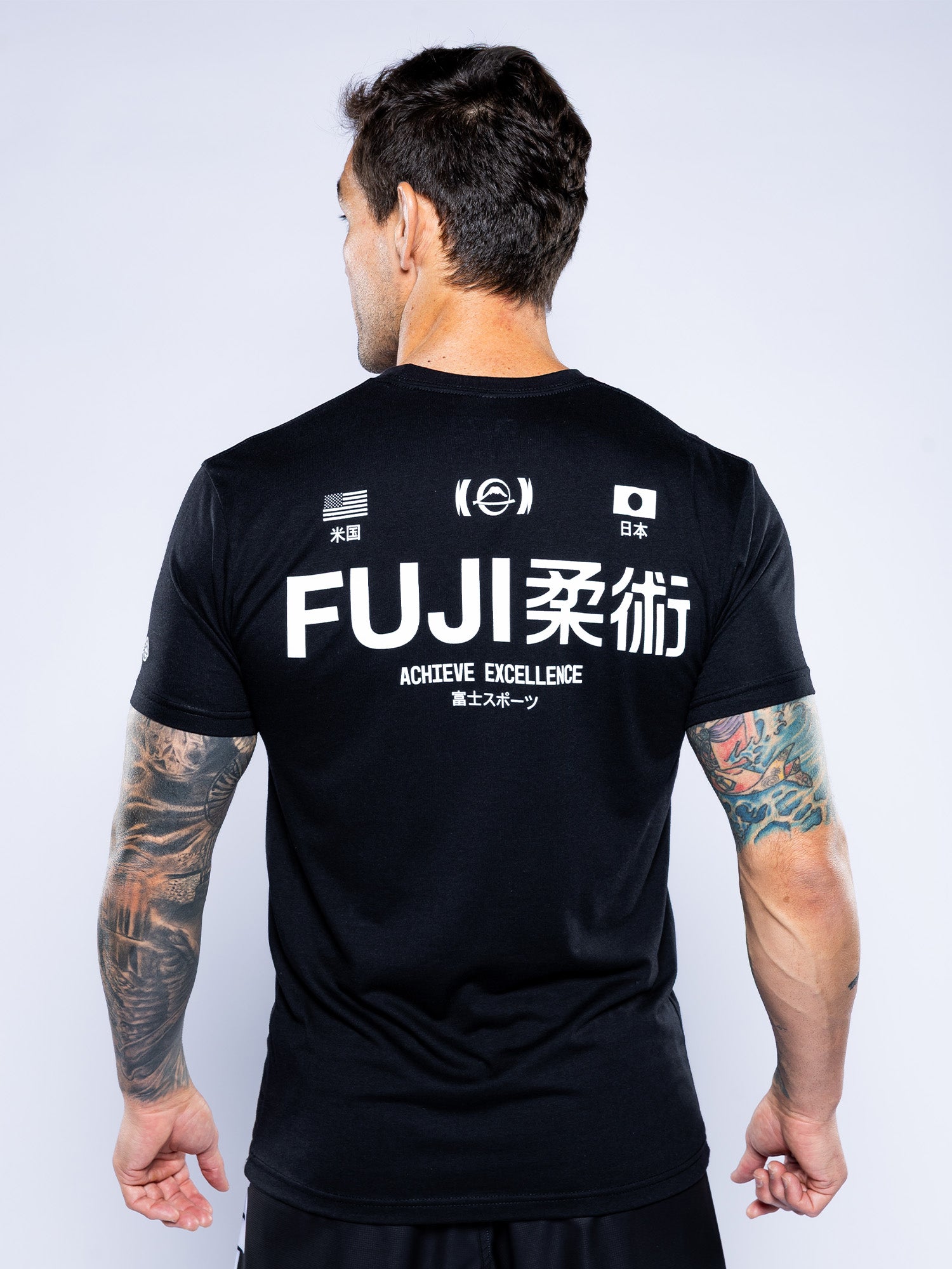 A tattooed man is seen from behind wearing the Iconic Jiu Jitsu Shirt Black featuring "FUJI 柔術 ACHIEVE EXCELLENCE" with US and Japanese flags and white graphics on the back.