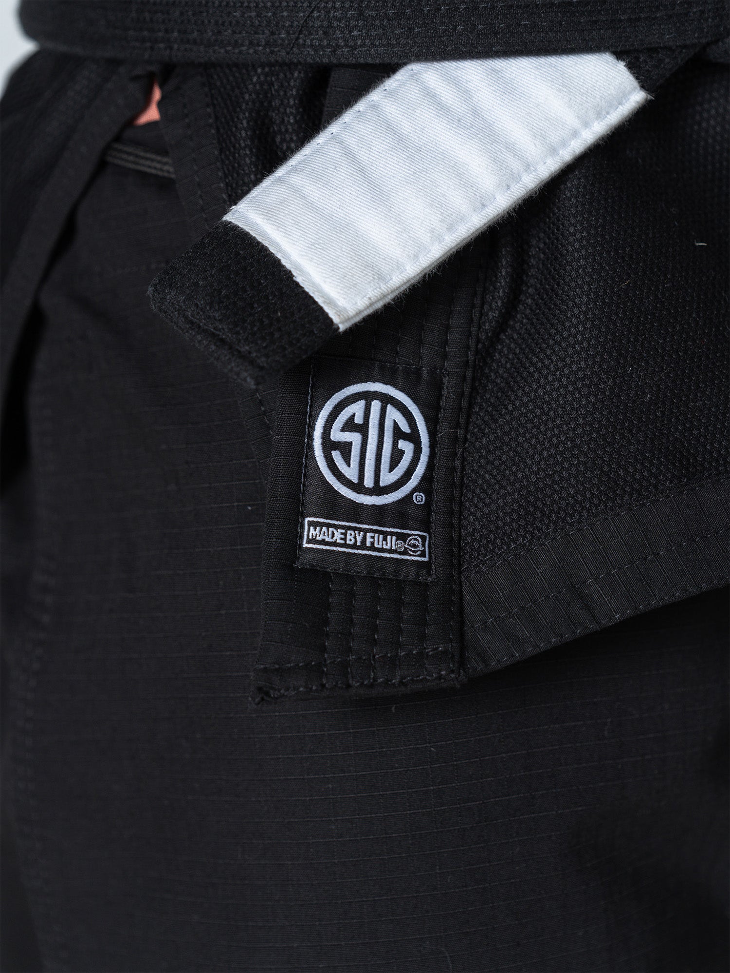 Close-up of the SIG SAUER Suparaito Gi Black, featuring a textured black fabric and a white-striped black belt. A SIG logo patch and "MADE BY FUJI" text appear on the label, with the belt tied at the waist.
