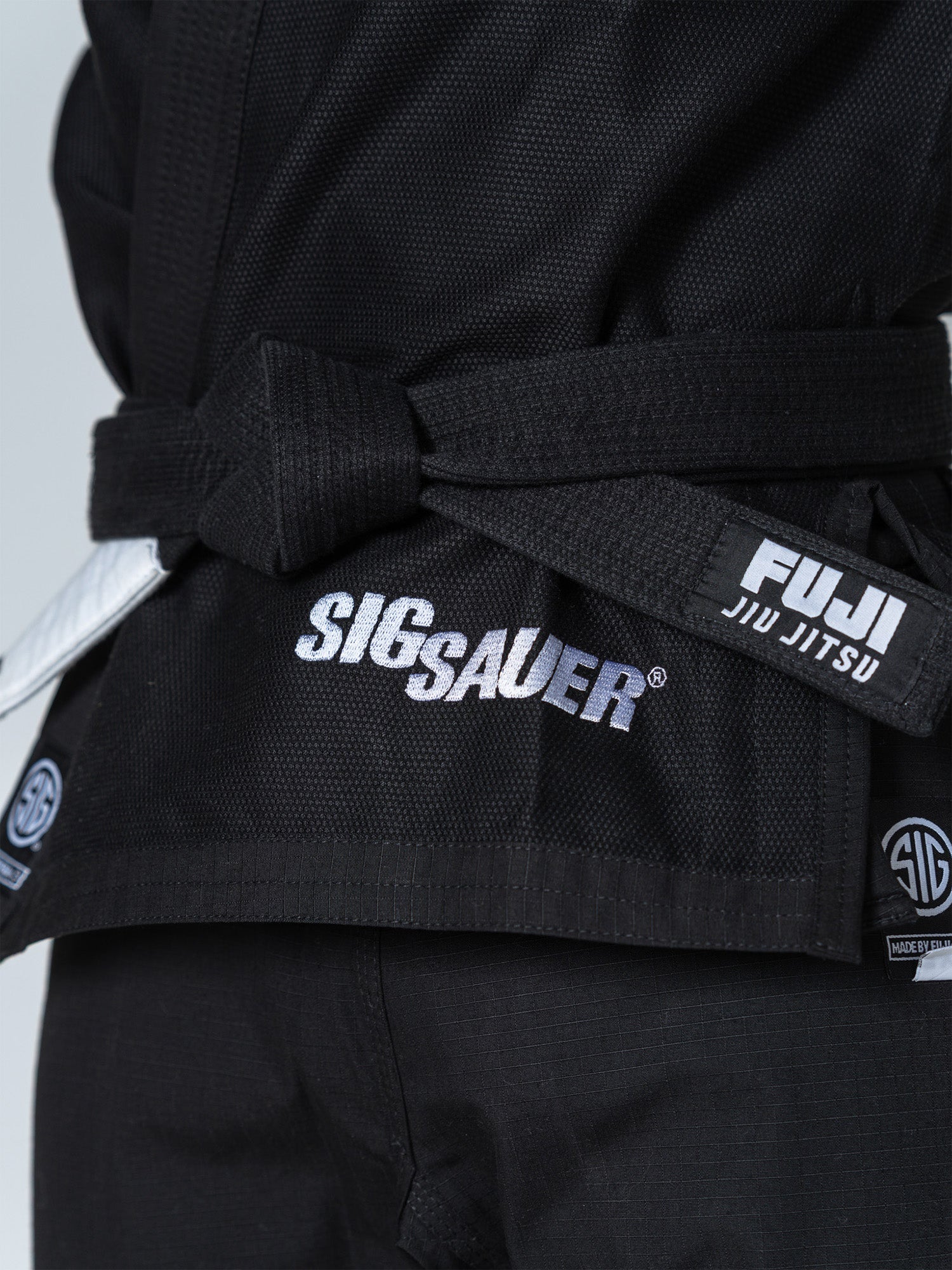 Close-up of a person wearing the SIG SAUER Suparaito Gi Black, featuring embroidered SIG SAUER and FUJI JIU JITSU logos on the jacket and belt, paired with a black belt.