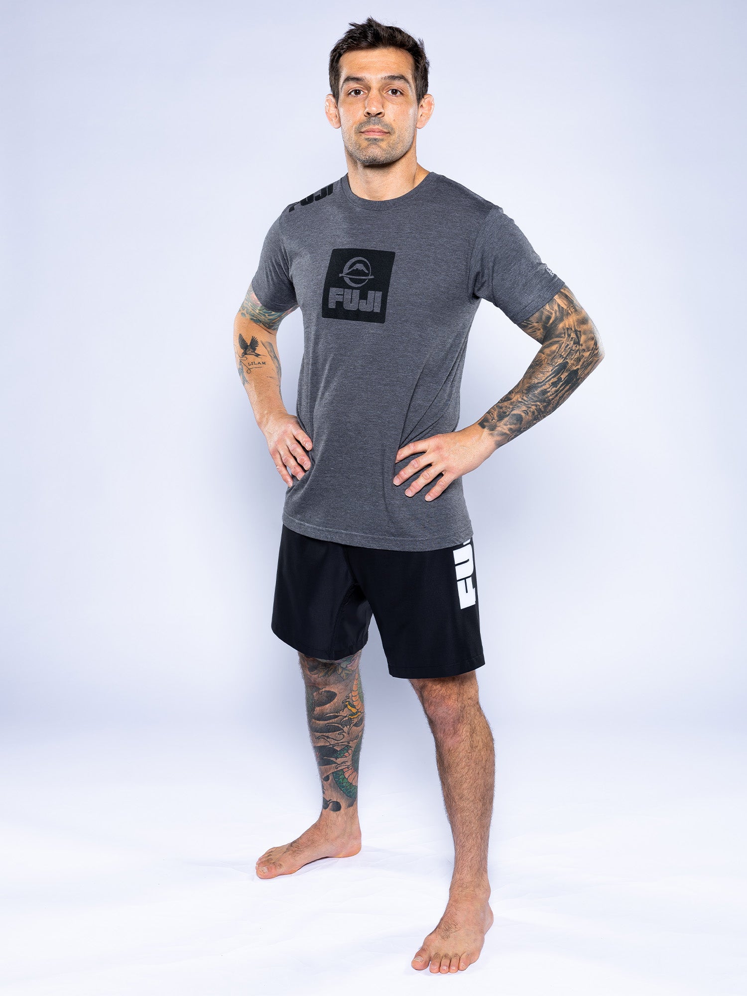 A man with tattooed arms and legs stands barefoot on a white background, wearing the Elevate Your Journey Shirt Charcoal and black FUJI shorts, with his hands on his hips and a neutral expression.