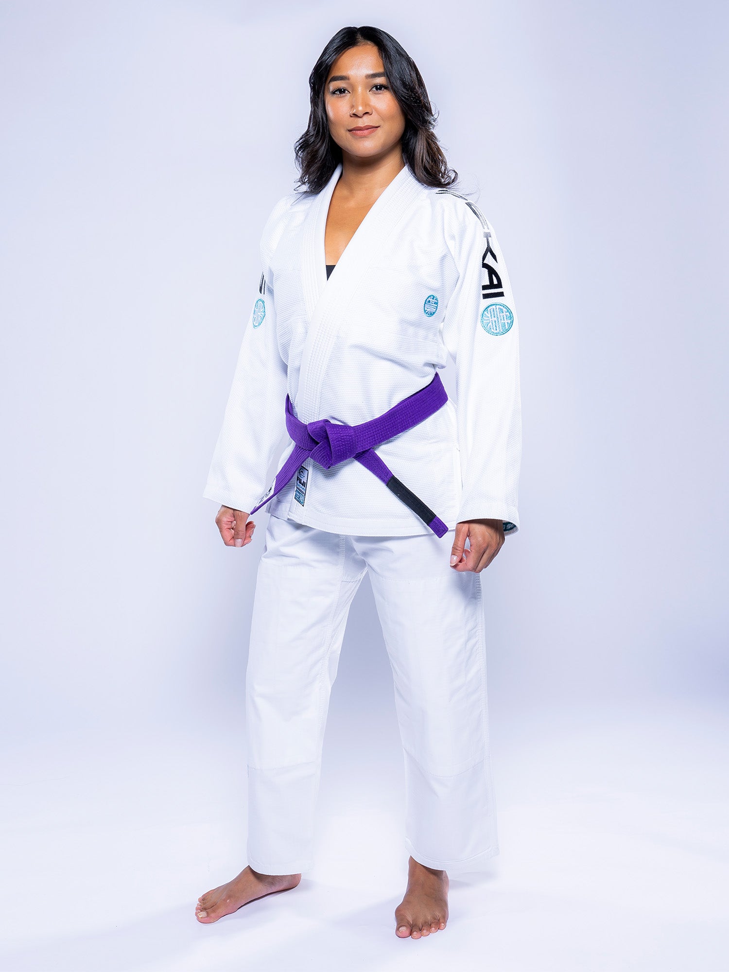 A woman with long dark hair stands confidently barefoot on a plain white background, wearing the Womens SEKAI 3.0 BJJ Gi White Teal and a purple belt, looking directly at the camera.