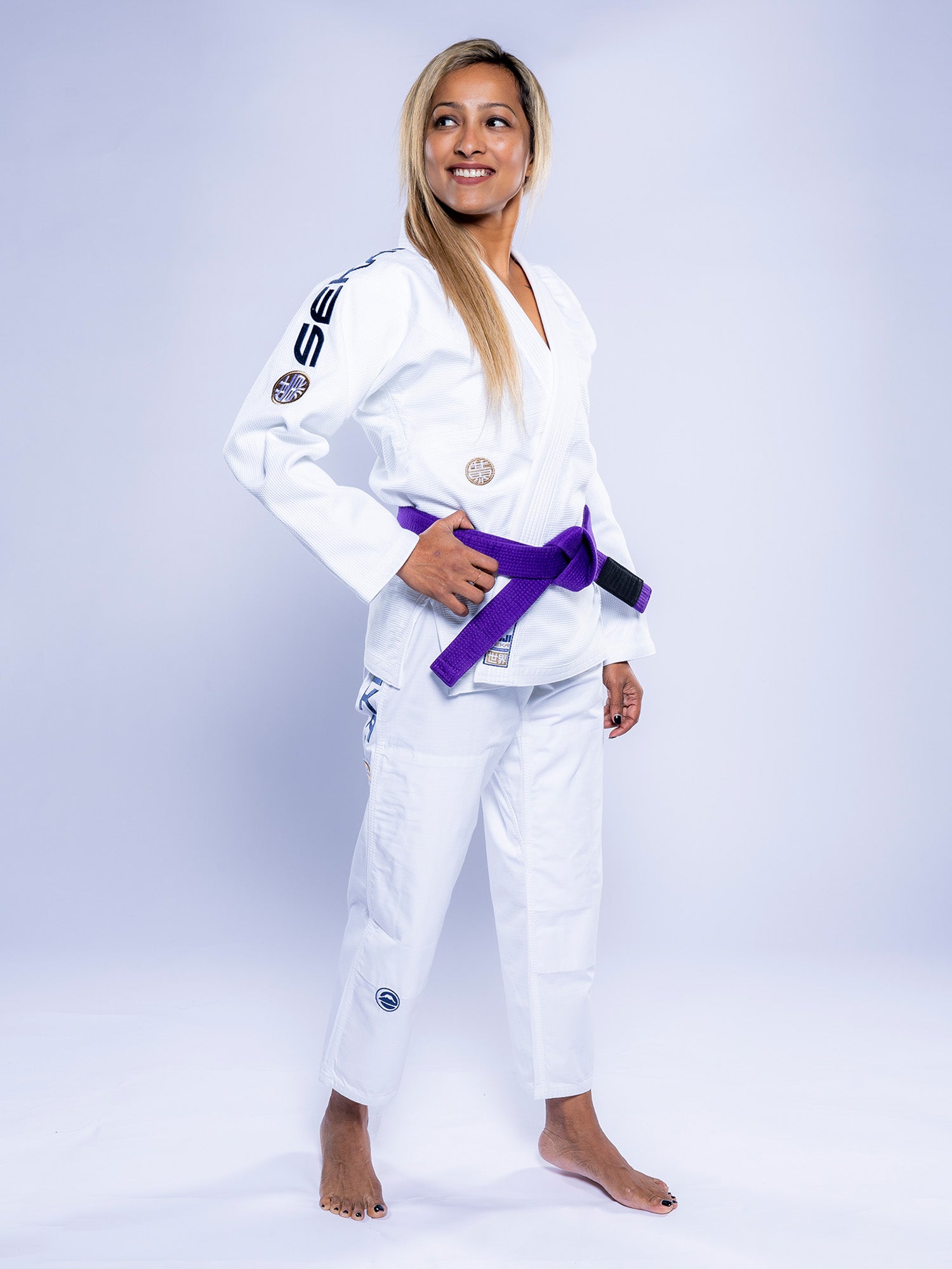 A woman stands barefoot on a white background, smiling and looking to the side while wearing the Womens SEKAI 3.0 BJJ Gi White Navy with a purple belt, her hands resting near her belt.