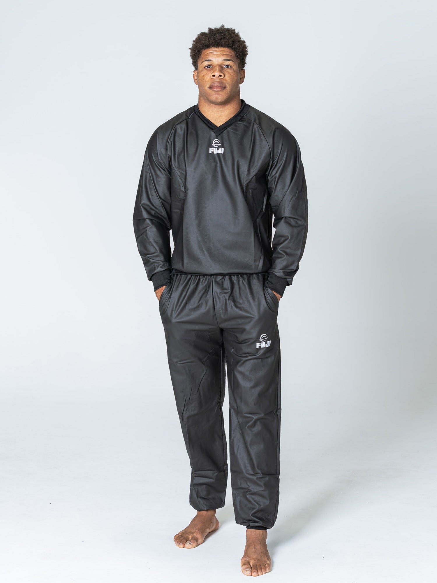 A person stands barefoot on a light background, wearing the Grappling Sauna Suit Black, a matching long-sleeve tracksuit set with FUJI logos on the chest and pants.