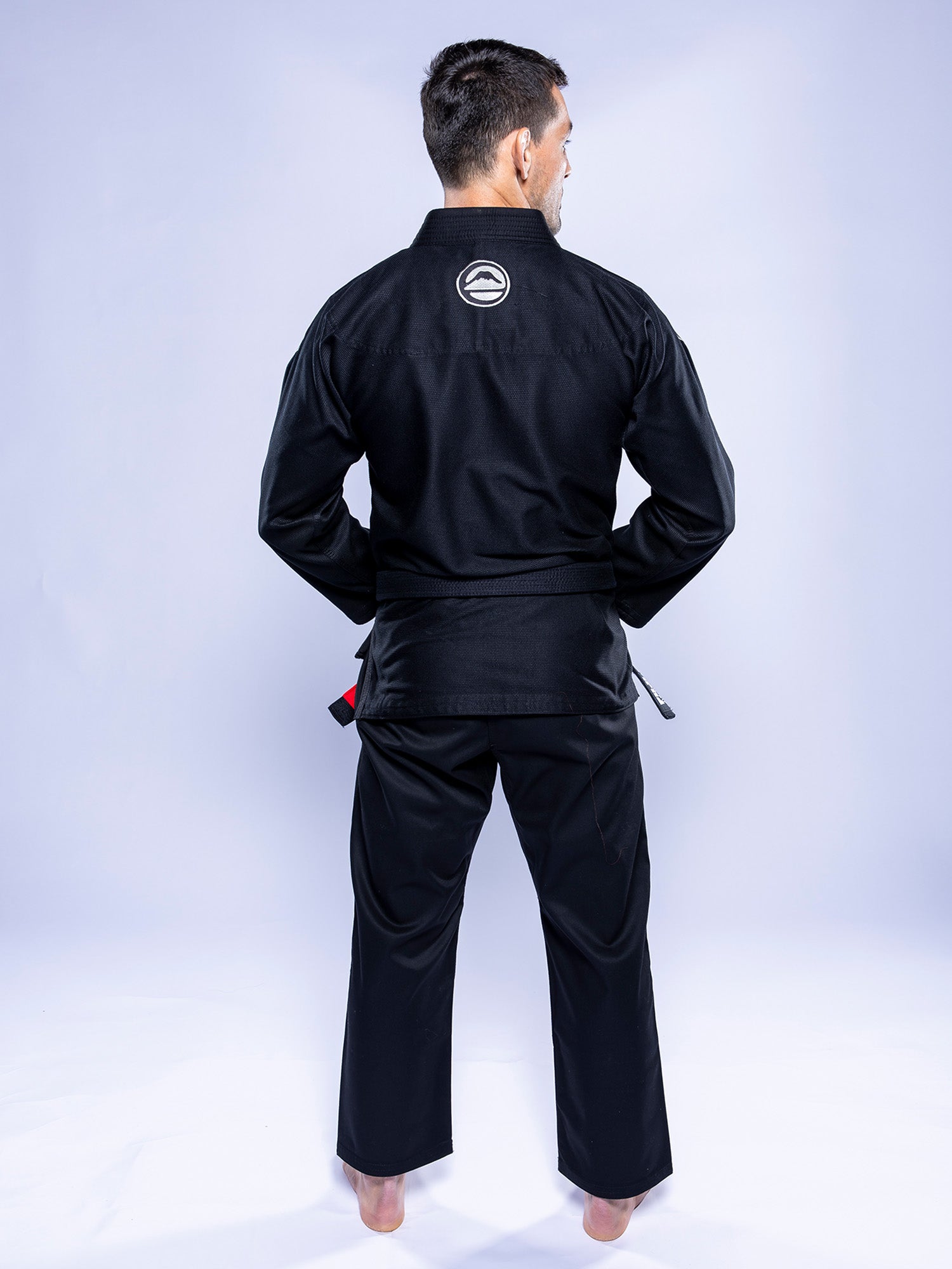 A person stands barefoot on a white surface, facing away from the camera, wearing the Baseline 2.0 BJJ Gi Black Grey with a circular logo on the upper back, against a plain light gray background.