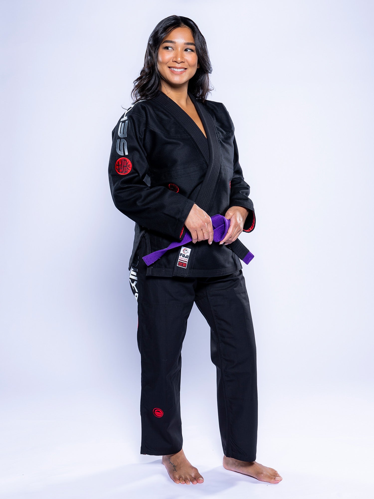 A woman stands barefoot on a white background, smiling and holding a purple belt. She is wearing the Womens SEKAI 3.0 BJJ Gi Black Grey, featuring red and white patches on the sleeves, pants, and chest.
