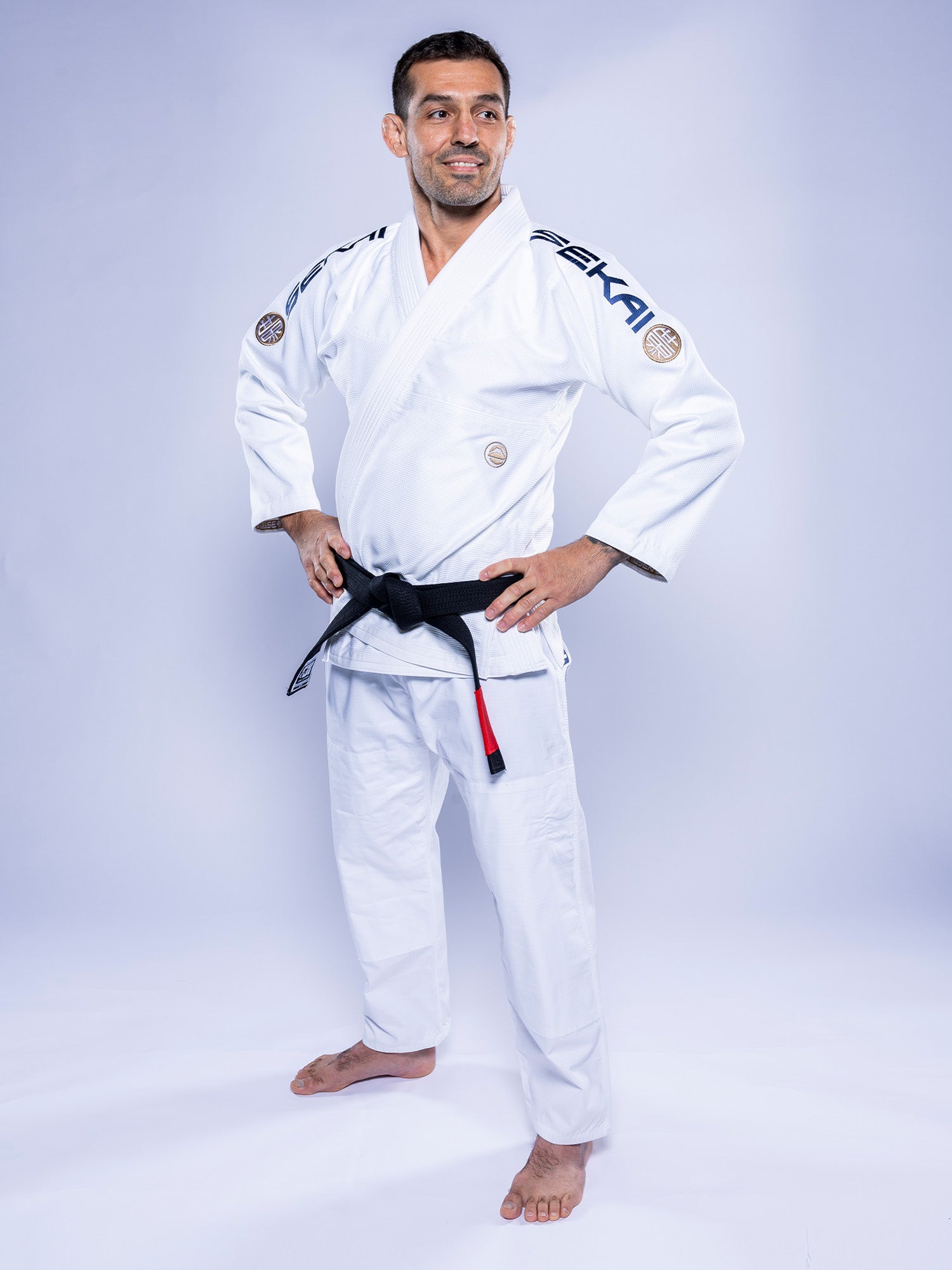 A man stands barefoot on a plain light background, smiling with his hands on his hips and wearing the SEKAI 3.0 BJJ Gi White Navy with a black belt.