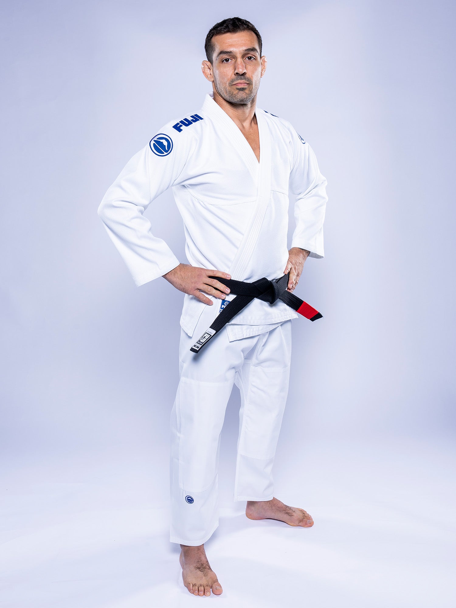 A man wears the Baseline 2.0 BJJ Gi White Navy with a black belt, stands barefoot on a white background, looking at the camera with his hands on his hips.