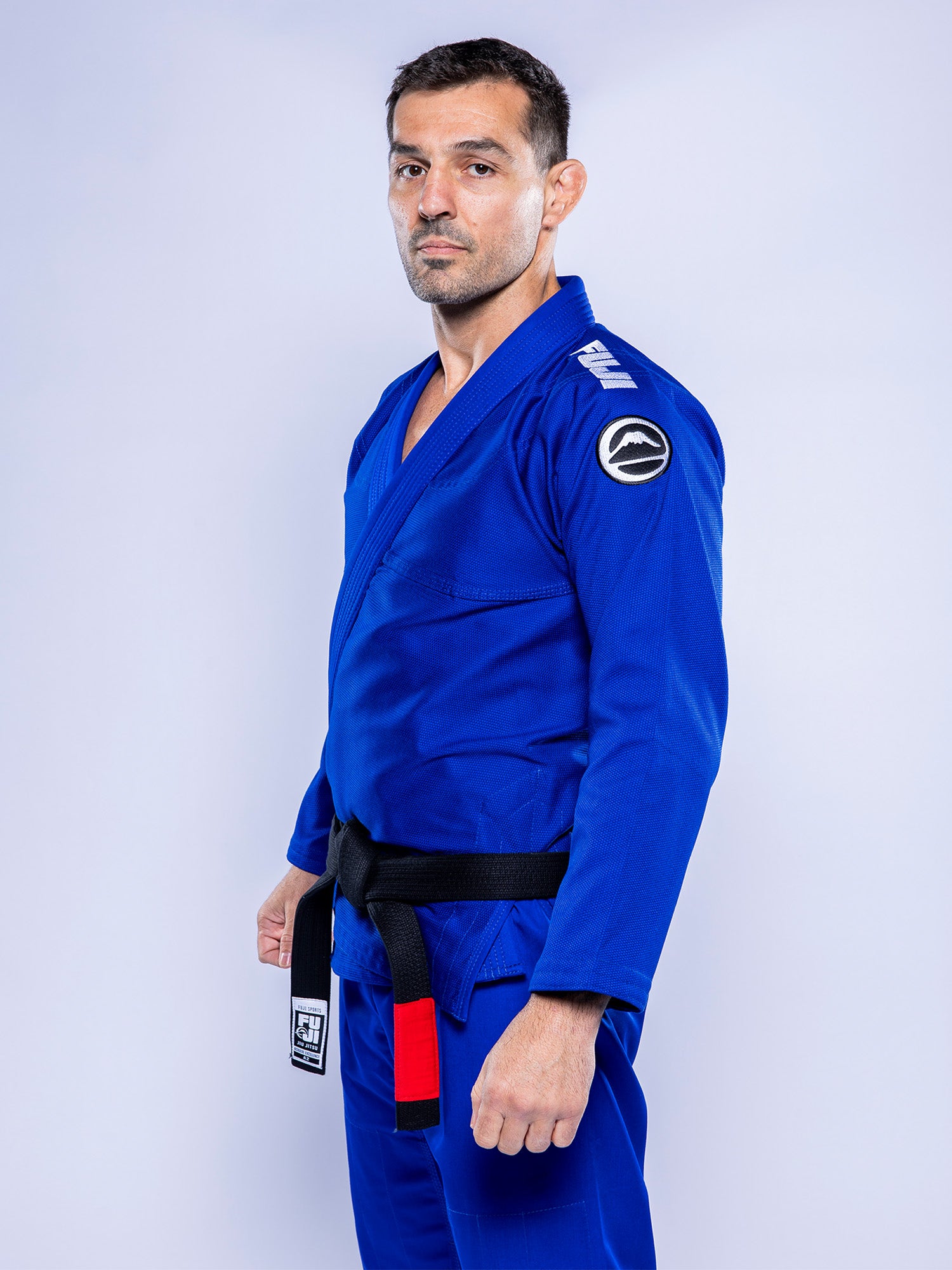 A man wears the Baseline 2.0 BJJ Gi Blue White with a black belt, standing slightly sideways and looking at the camera against a plain light background.