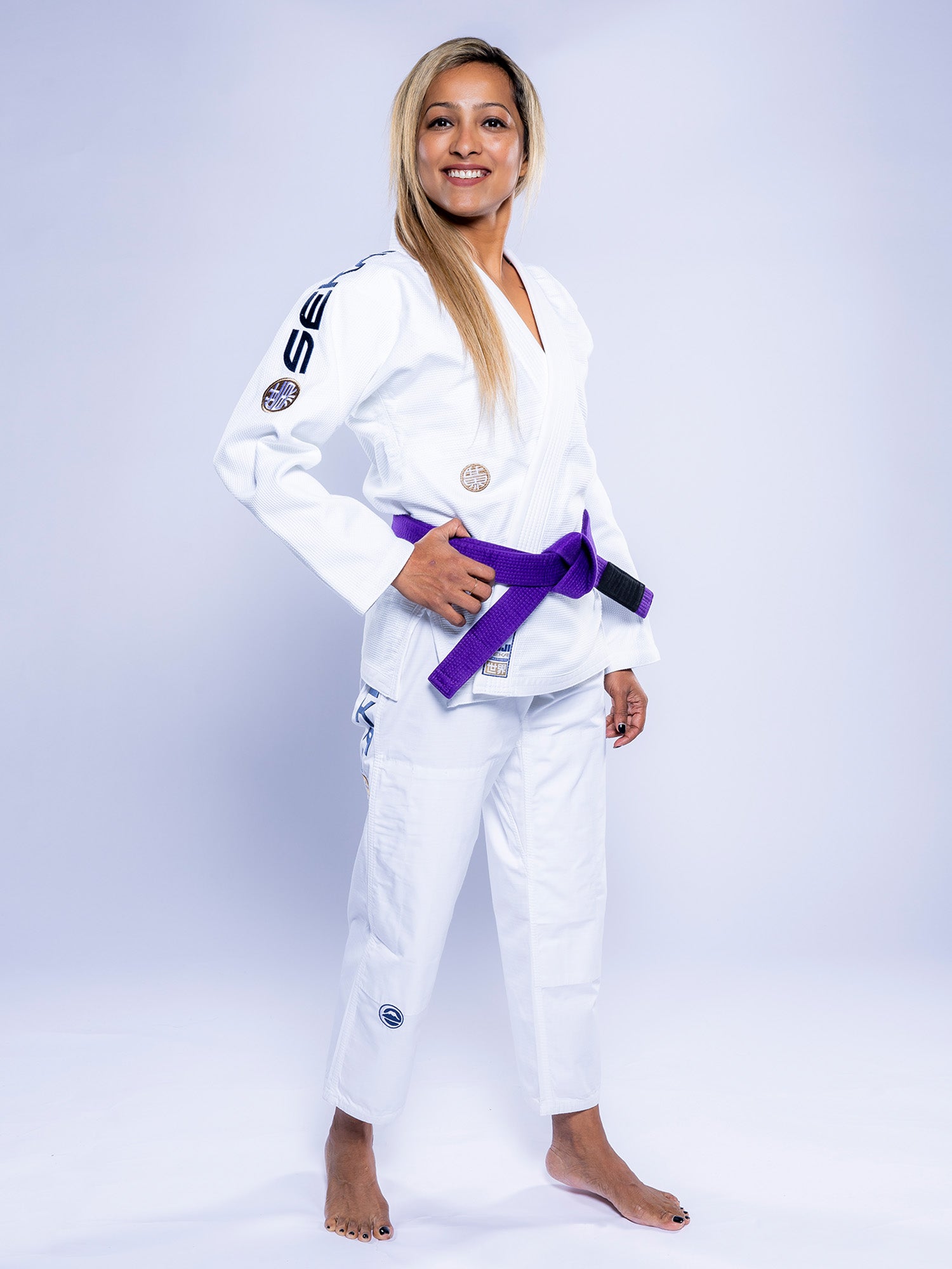 A woman smiles barefoot on a white backdrop, wearing the Womens SEKAI 3.0 BJJ Gi in white/navy with a purple belt tied around her waist.