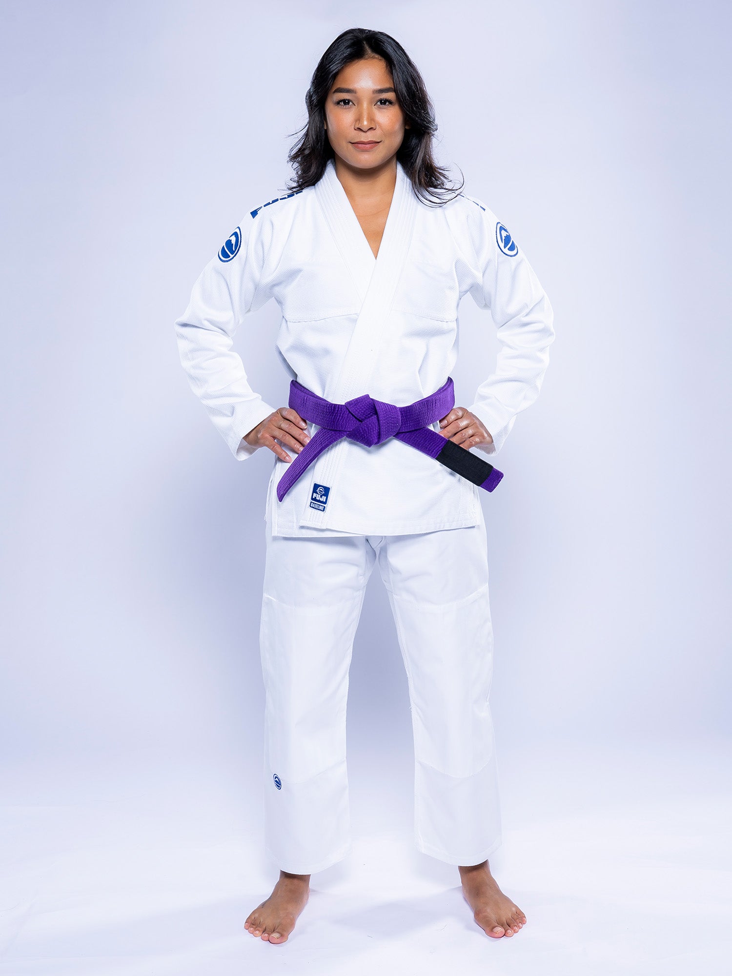 A woman stands barefoot on a white background, confidently wearing the Womens Baseline 2.0 BJJ Gi White Navy with a purple belt, hands on her hips and looking at the camera.