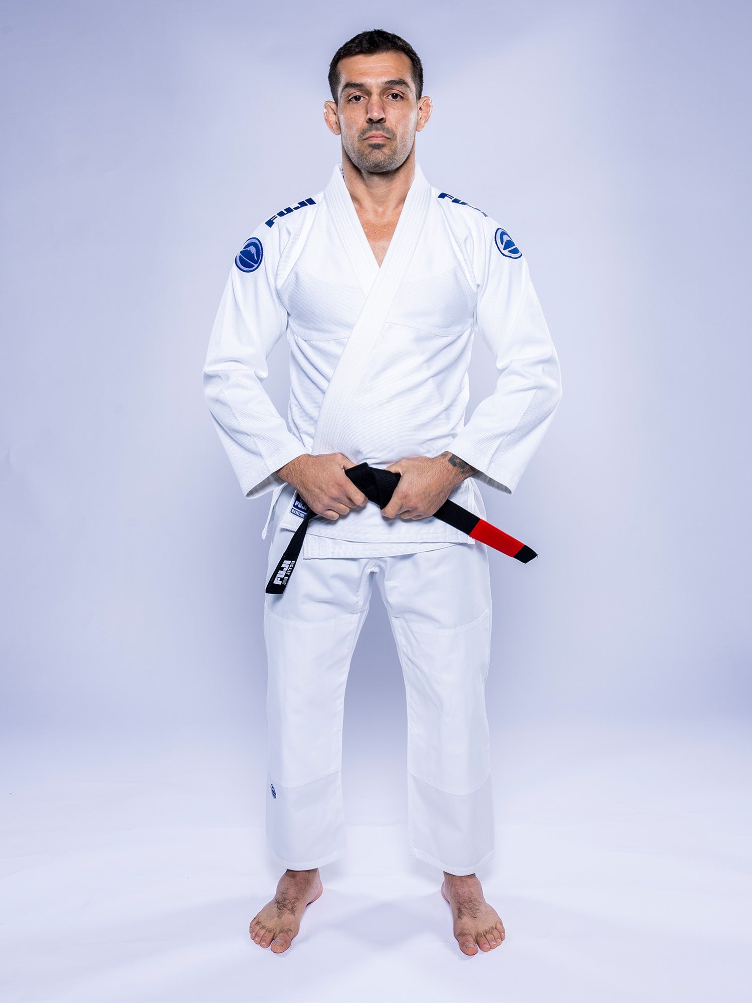 A man wearing the Baseline 2.0 BJJ Gi White Navy stands barefoot on a white background, holding a black belt with a red bar, facing forward with a neutral expression and his hands resting on the belt.