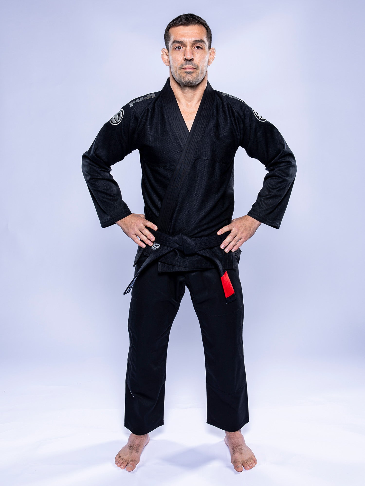 A man stands barefoot on a white surface, wearing the Baseline 2.0 BJJ Gi Black Grey with a red bar on the belt. His hands rest on his hips as he faces forward against a plain light background.