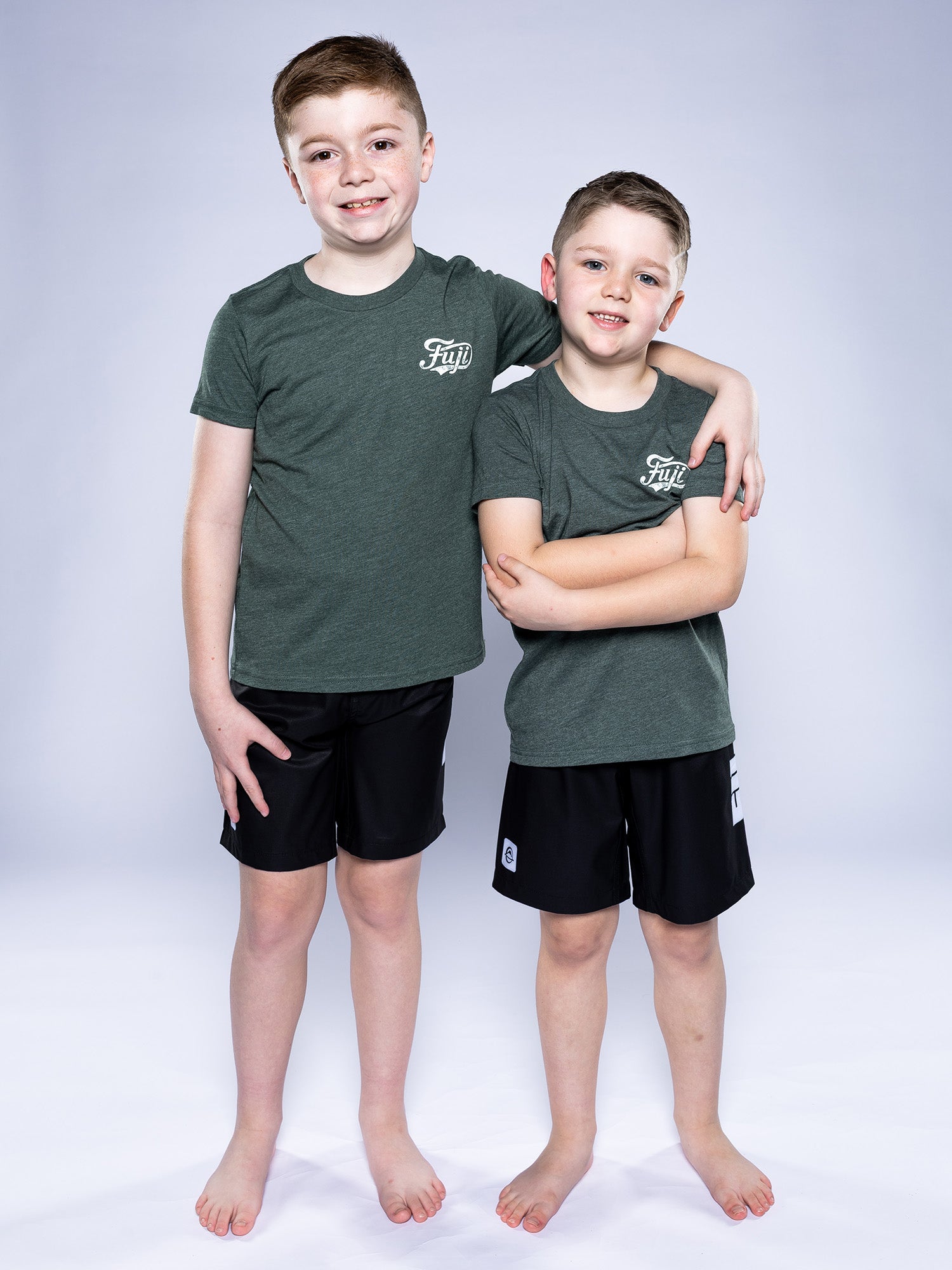 Two boys stand barefoot on a white backdrop, smiling. The older has his arm around the younger. Both wear matching Kids Jiu Jitsu Flow T-Shirts in green with white logos and black shorts.