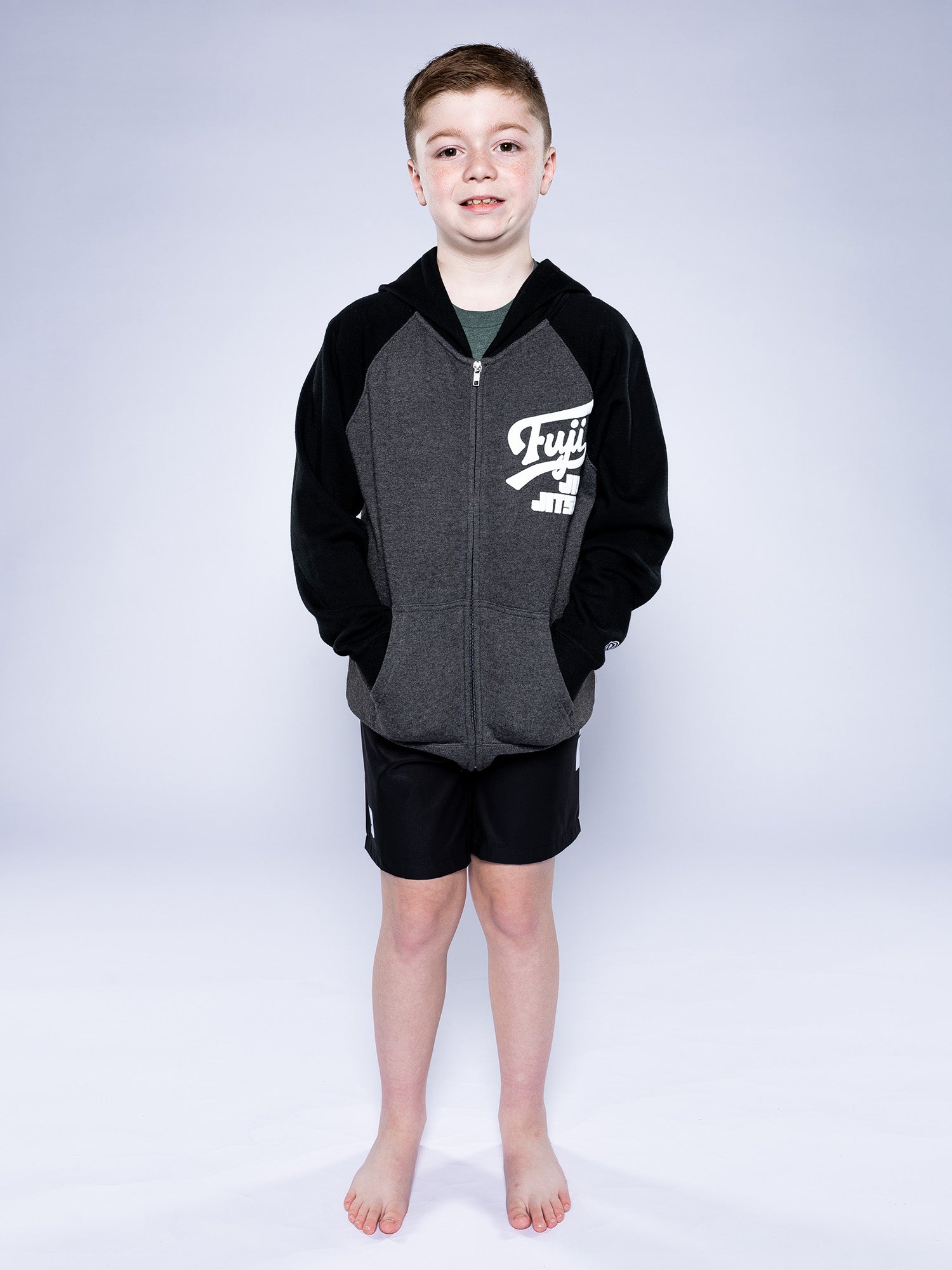 A young boy with short hair stands barefoot on a white backdrop, wearing the Kids Jiu Jitsu Varsity Zip Hoodie Charcoal with black sleeves and black athletic shorts. He smiles slightly and looks at the camera.
