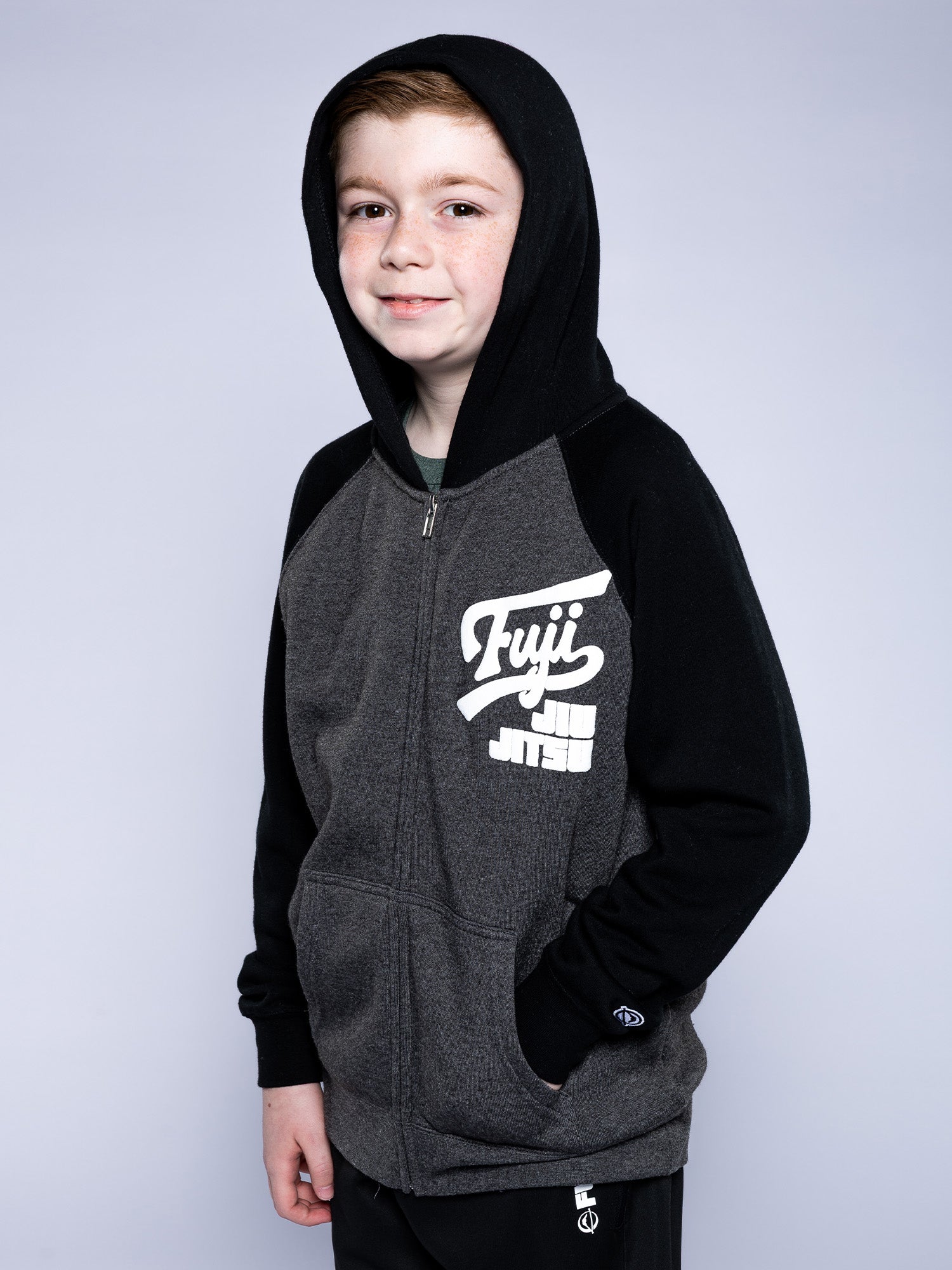 A young boy smiles at the camera with one hand in his pocket, wearing the Kids Jiu Jitsu Varsity Zip Hoodie in charcoal against a light gray background.