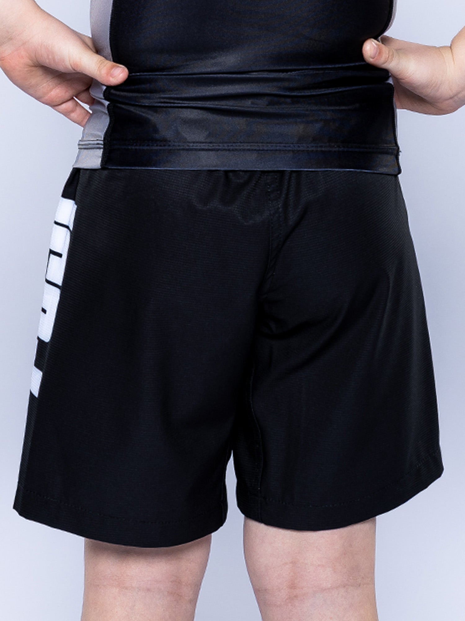 A child wearing the Baseline Grappling Kids Shorts in black and gray stands with hands on hips, facing away from the camera. The shorts feature a white graphic detail on the side.