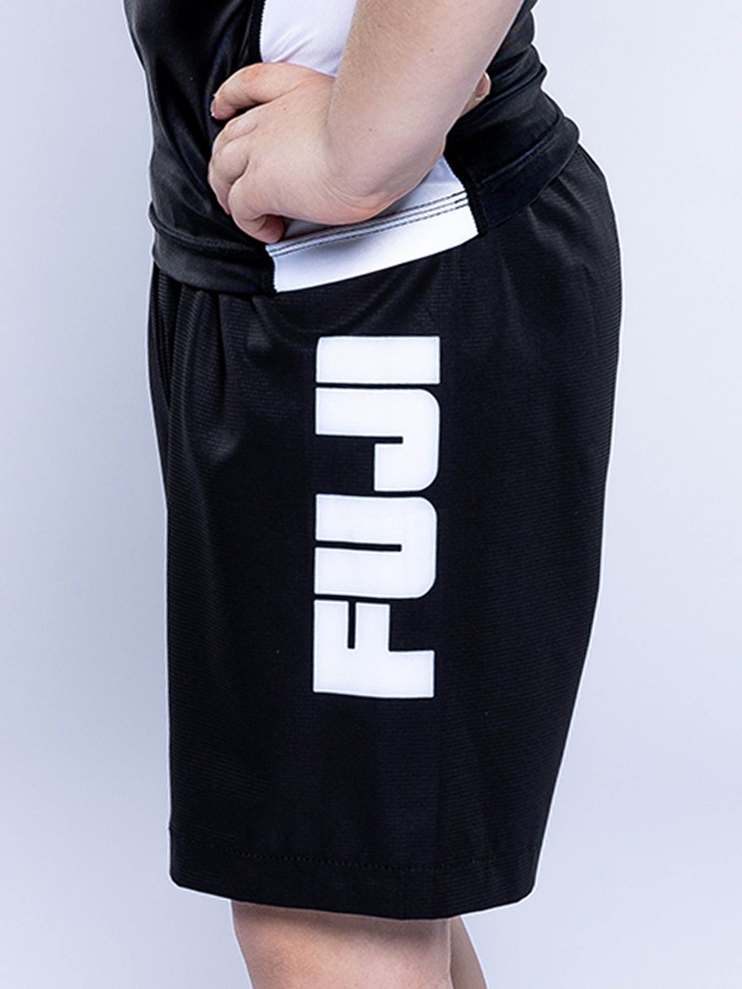 A person wearing Baseline Grappling Kids Shorts in black with "FUJI" in large white letters vertically on the side; only their lower torso and upper legs are visible.