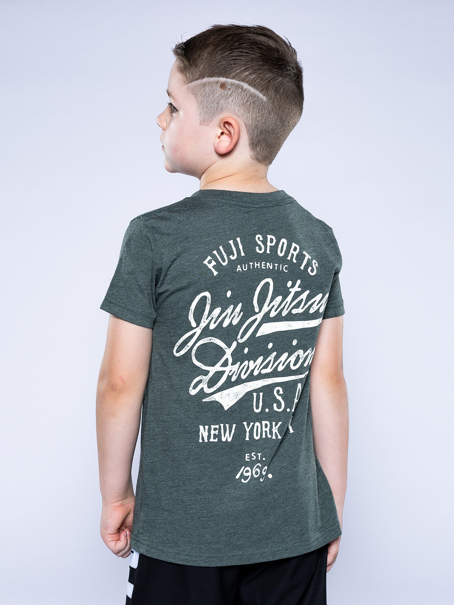 A young boy with short brown hair and shaved lines stands facing away, wearing the Kids Jiu Jitsu Flow T-Shirt Green, which features white text: “Fuji Sports Jiu Jitsu Division U.S.A. New York Est. 1969.”.