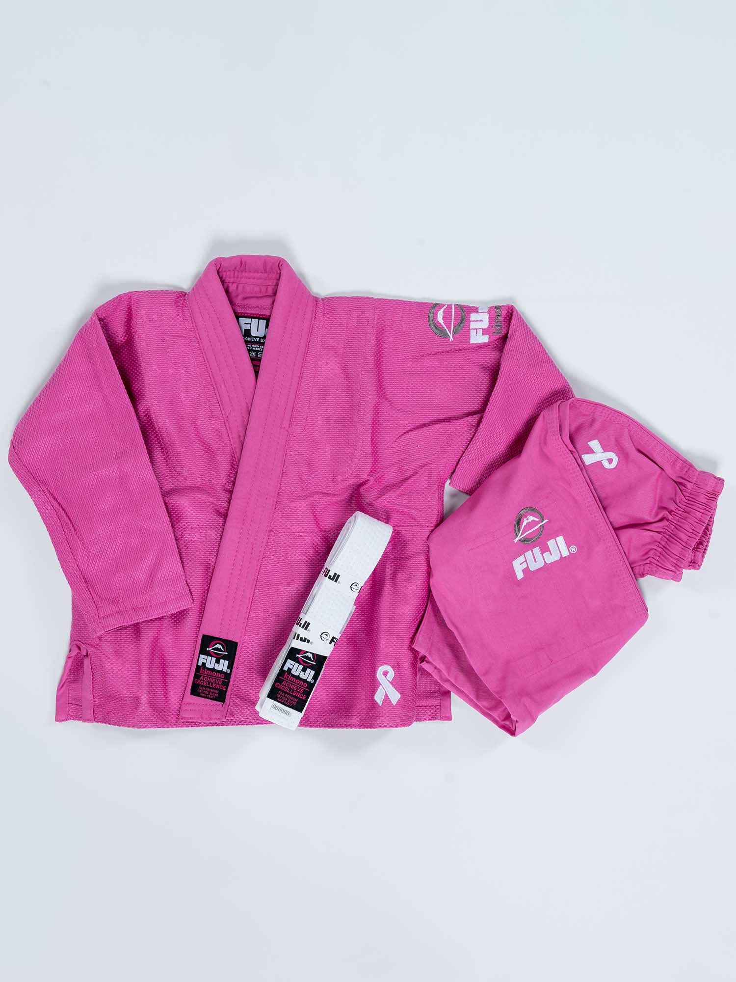 A pink FUJI Baby Judo Gi set, including a jacket, pants, and a white belt with black stripes, is laid out on a white background. The gi features white ribbon symbols supporting breast cancer awareness.