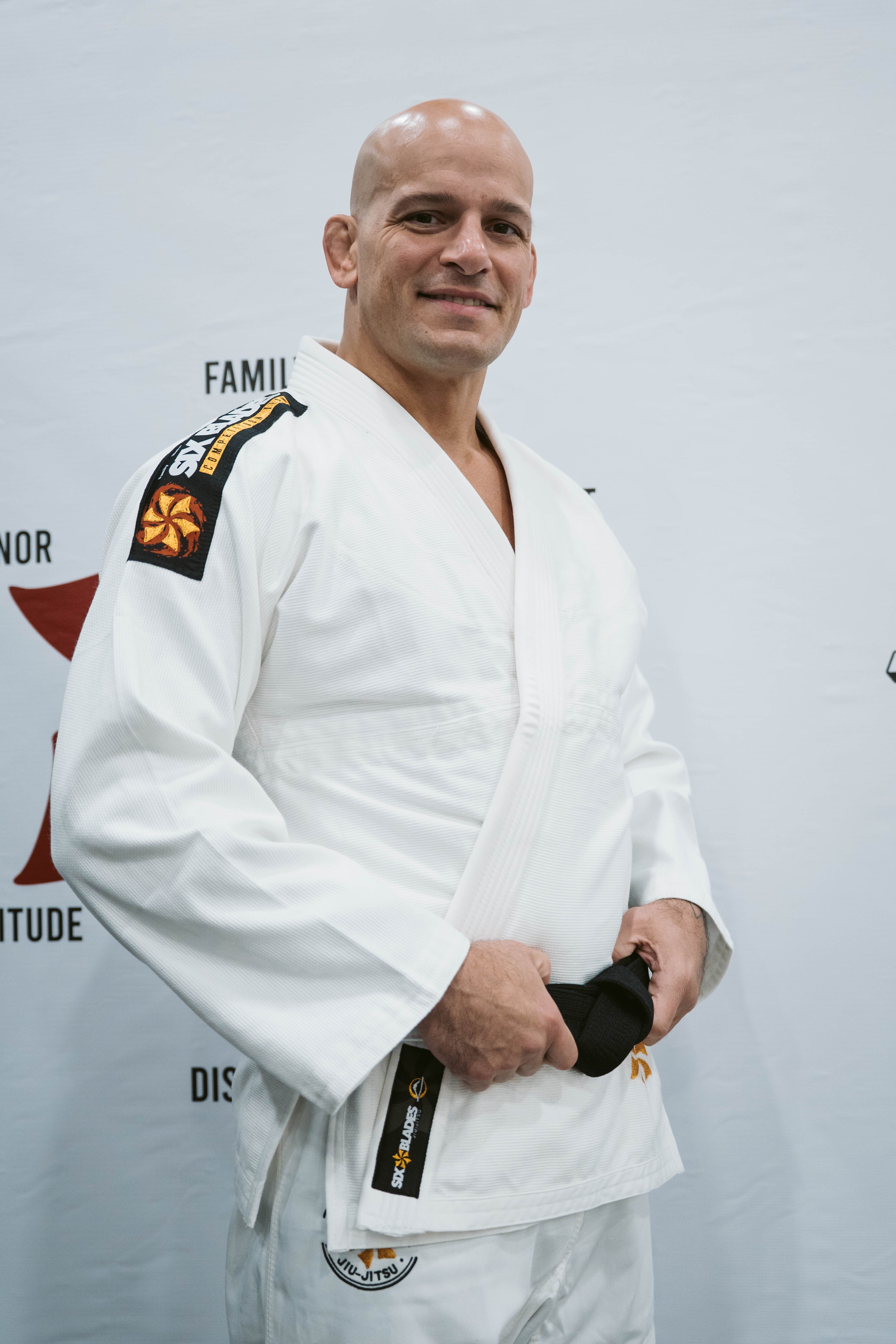 A man in a white Six Blades Competition Gi stands smiling, holding a black belt in front of a white backdrop with partially visible text and logos.