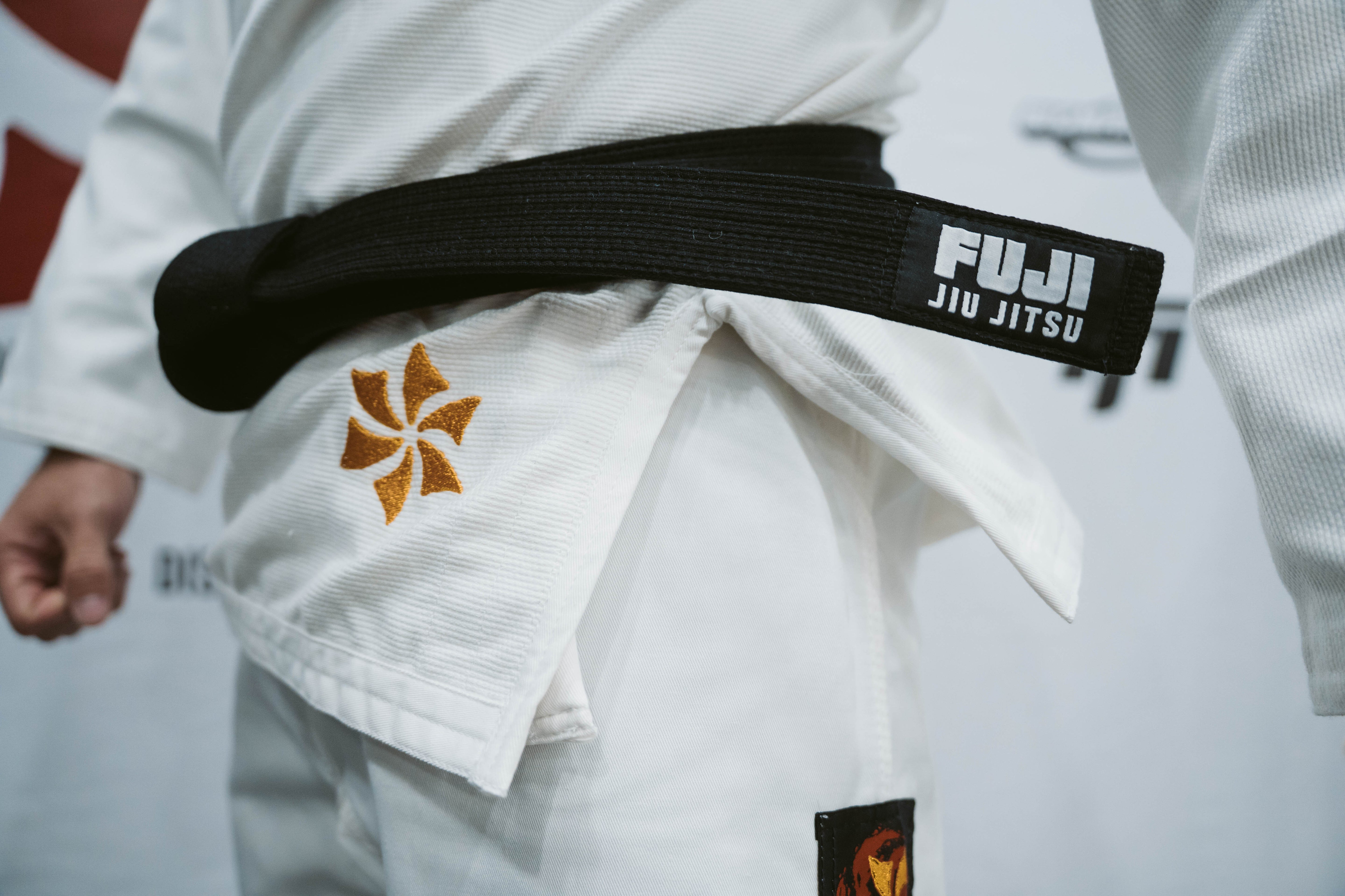 A close-up of a person wearing the Six Blades Competition Gi, a white jiu jitsu uniform with a black belt. The gi features embroidered patches, including an orange flower and a rectangular patch near the hip.