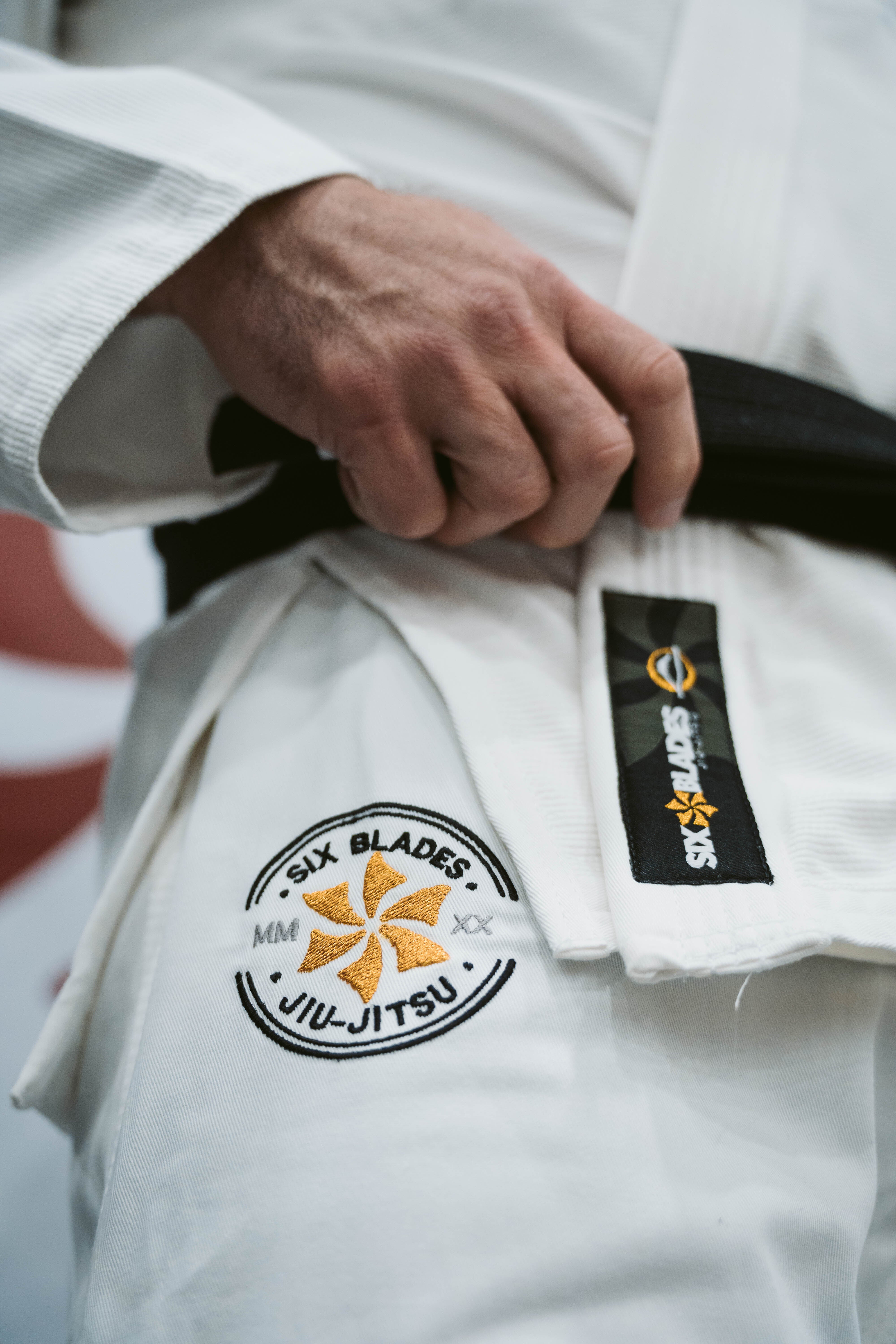 A person wearing the Six Blades Competition Gi holds a black belt, featuring a Six Blades Jiu-Jitsu patch on the gi and a Six Blades logo on the belt.