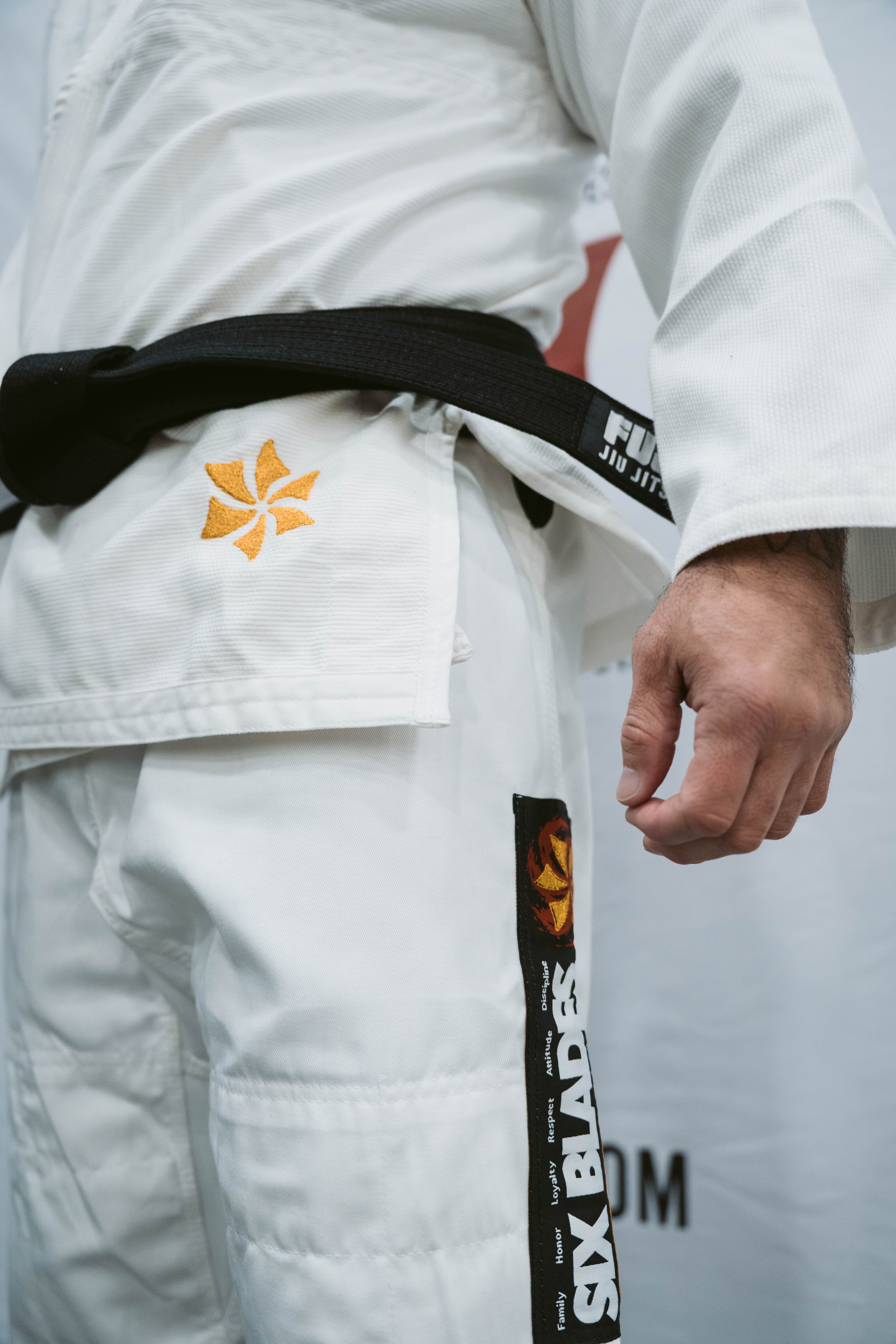 A person wearing the Six Blades Competition Gi, a white Brazilian Jiu-Jitsu gi with orange and black patches—including a Six Blades patch—and a black belt. Only their torso and right hand are shown.