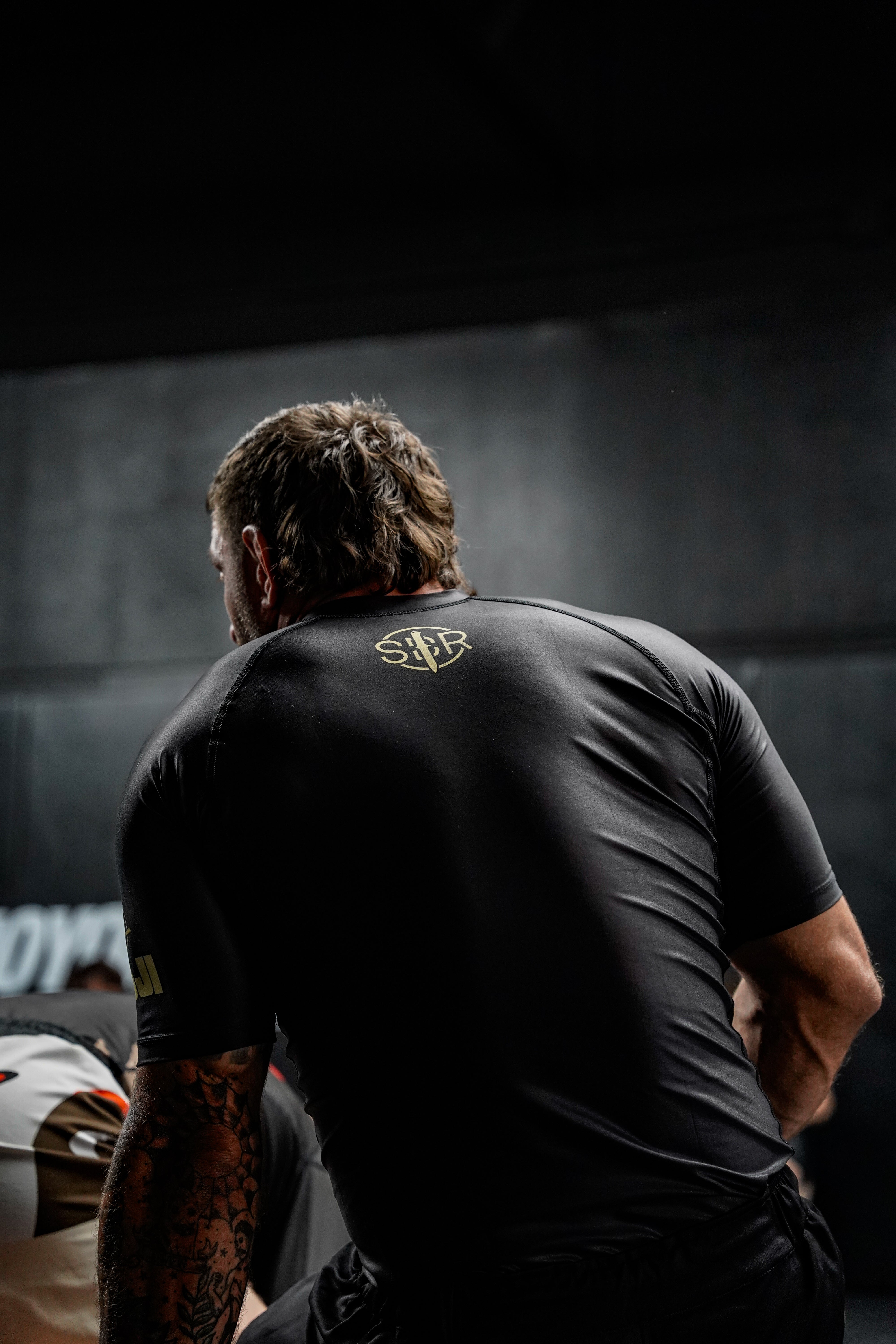 A man wears the Sheepdog Response Essential Short Sleeve Rashguard Black, leaning forward in a dark indoor gym with gold lettering visible on the back and another person partially seen beside him.