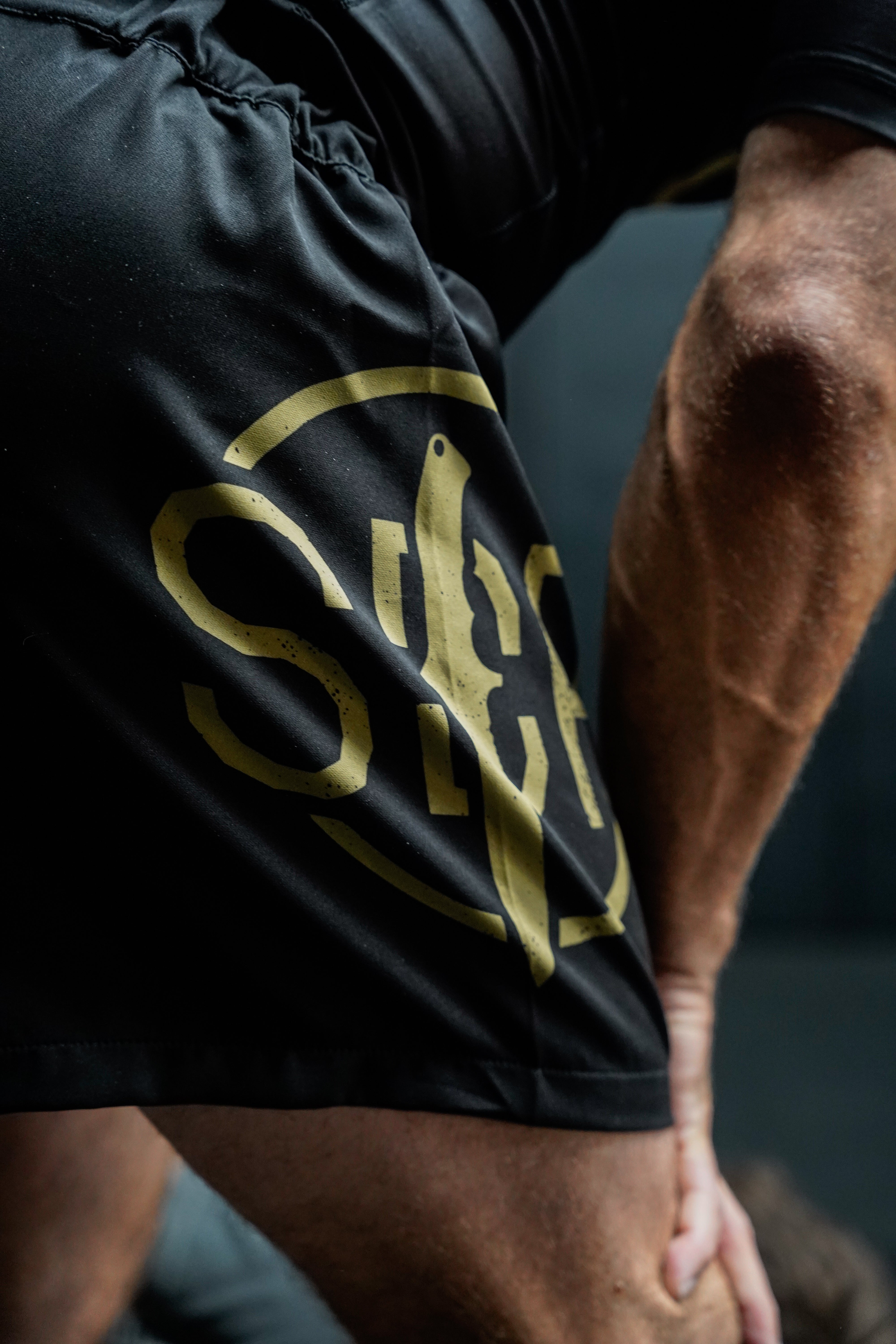 A close-up of a person wearing Sheepdog Response Essential Shorts Black, displaying the gold logo as they rest their hand on a bent knee during physical activity.
