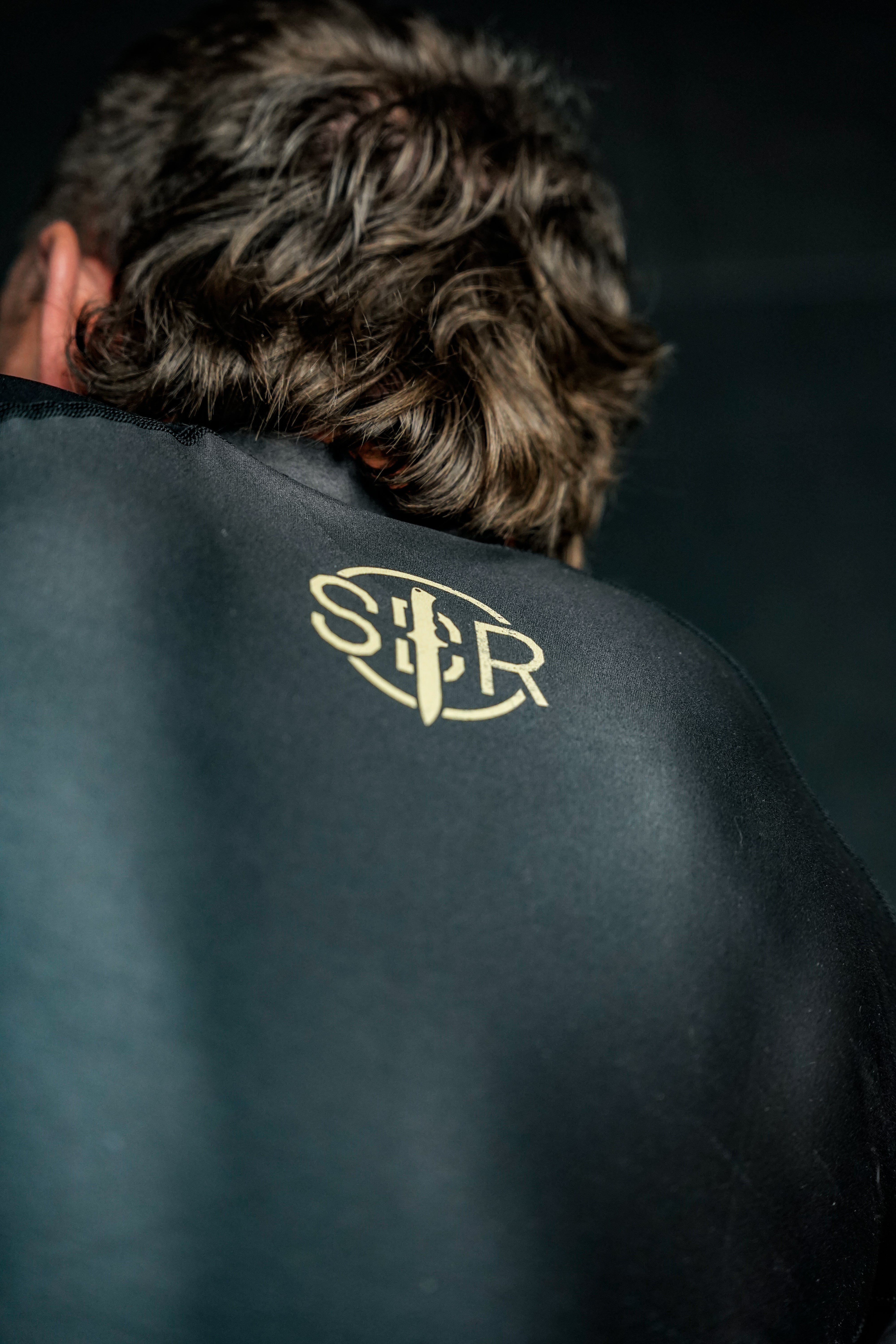 A person with brown, wavy hair is seen from behind, wearing the Sheepdog Response Essential Short Sleeve Rashguard Black, which features a gold SCR logo and vertical sword design.