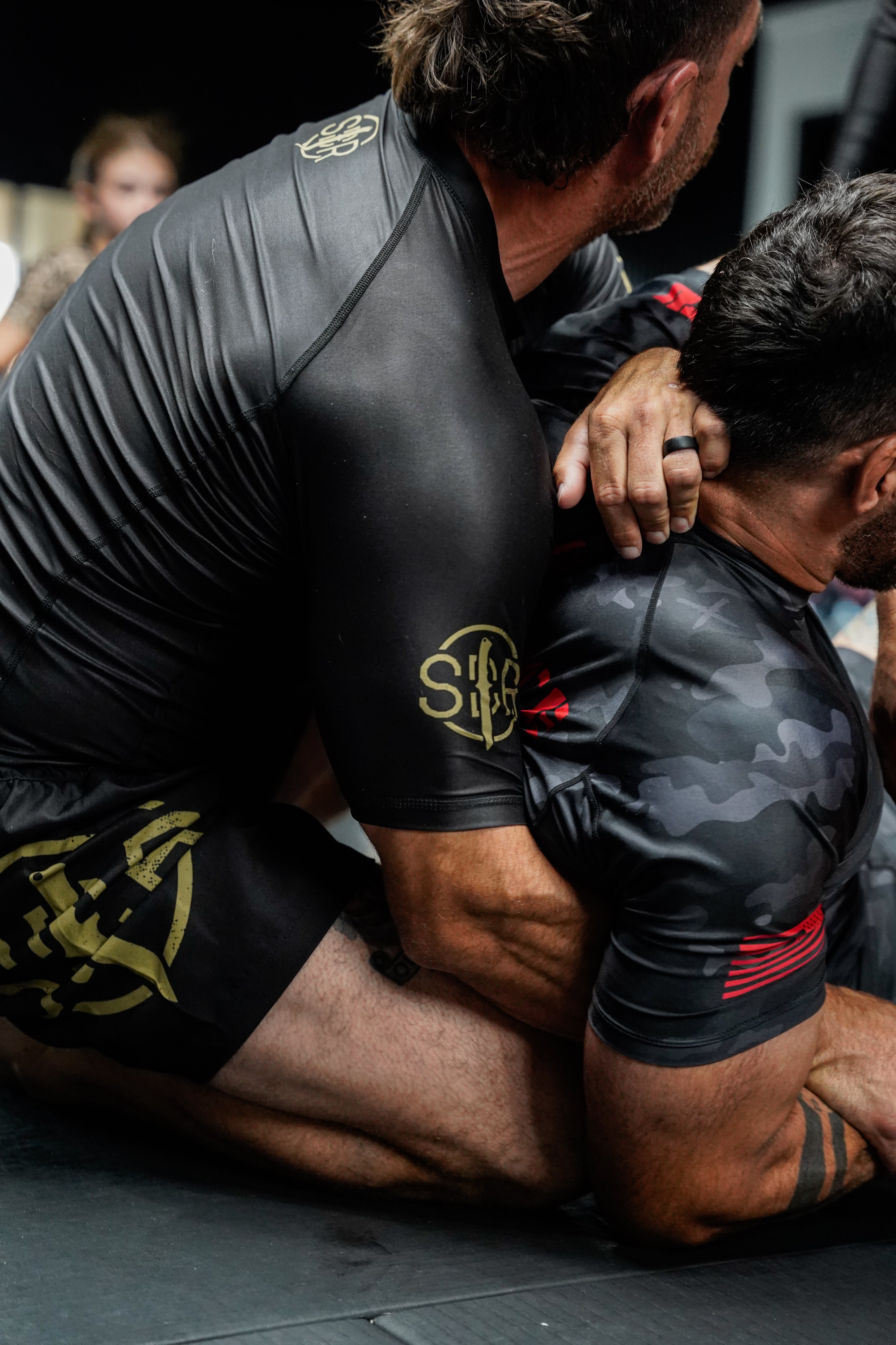 Two men wearing black athletic gear, including the Sheepdog Response Essential Short Sleeve Rashguard Black, grapple on a mat during a jiu-jitsu match. One man applies a hold from behind with his arms around the other's upper body.