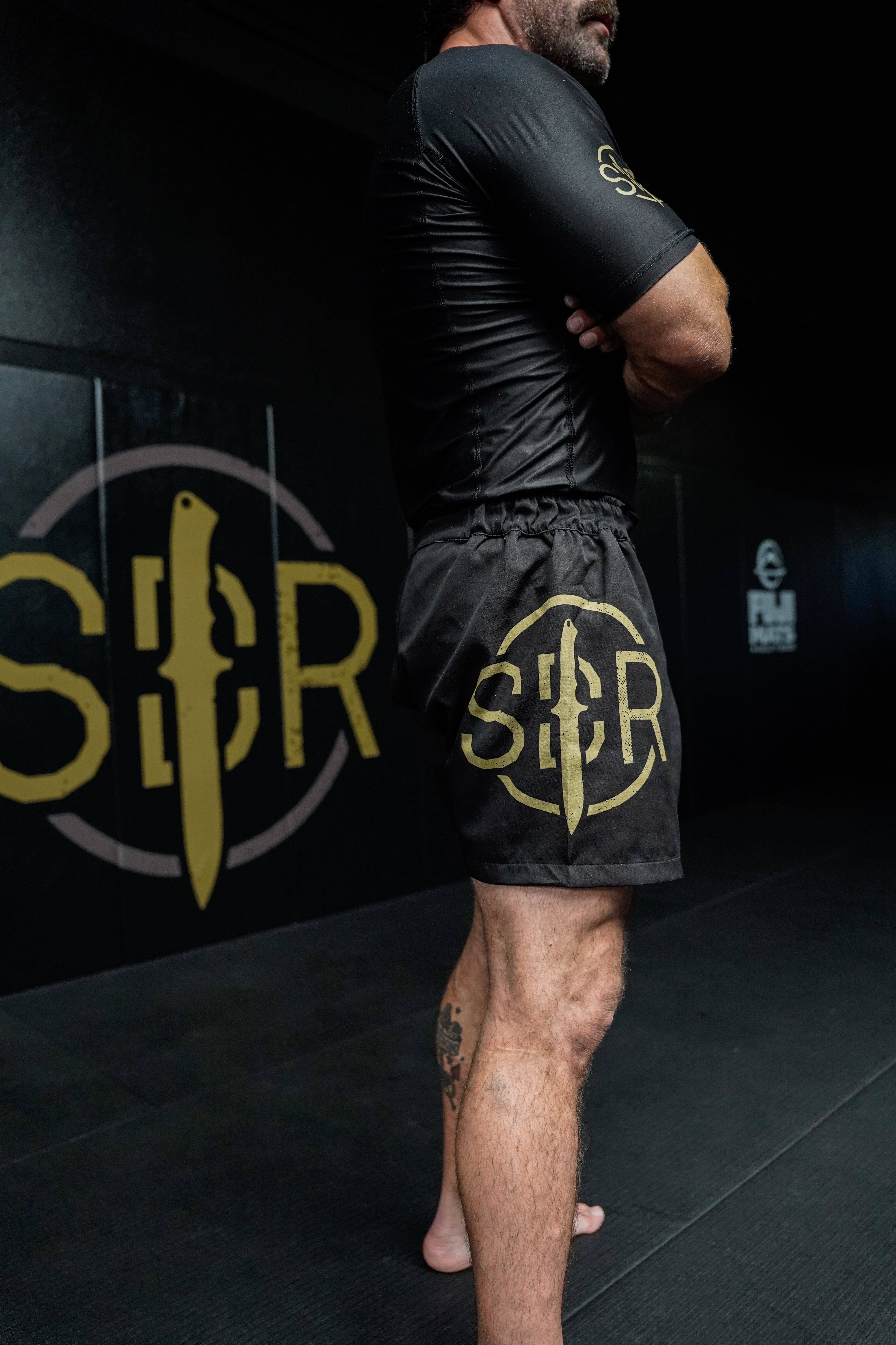 A man stands barefoot on a mat in a gym, arms crossed, wearing Sheepdog Response Essential Shorts Black with gold knife and SBR logos. Matching wall graphics are visible in the background.