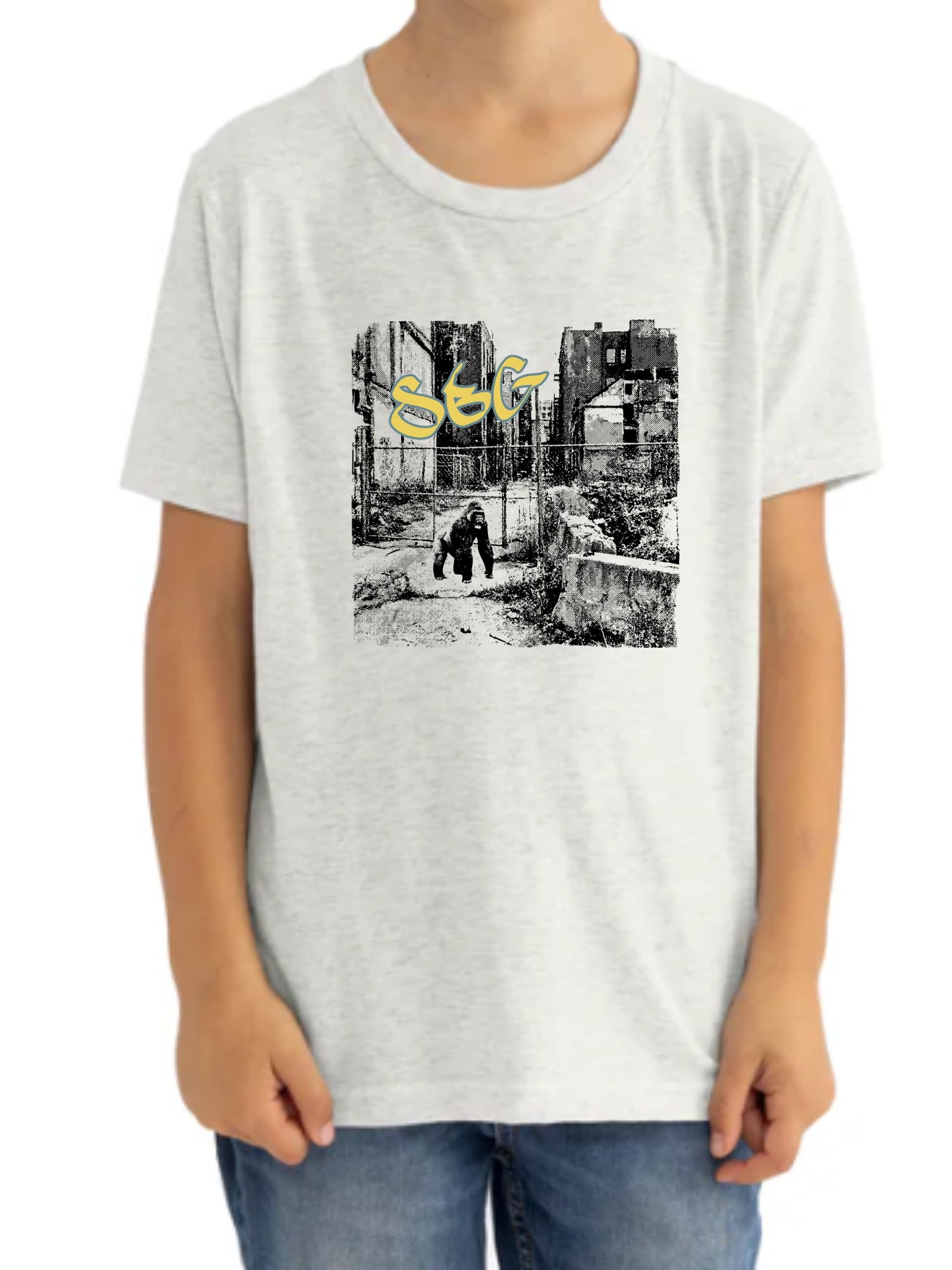 A child wears the SBG Junkyard Youth T-Shirt in Heather White, featuring a black-and-white urban scene graphic with a crouching figure and yellow graffiti-style "STG" text above.