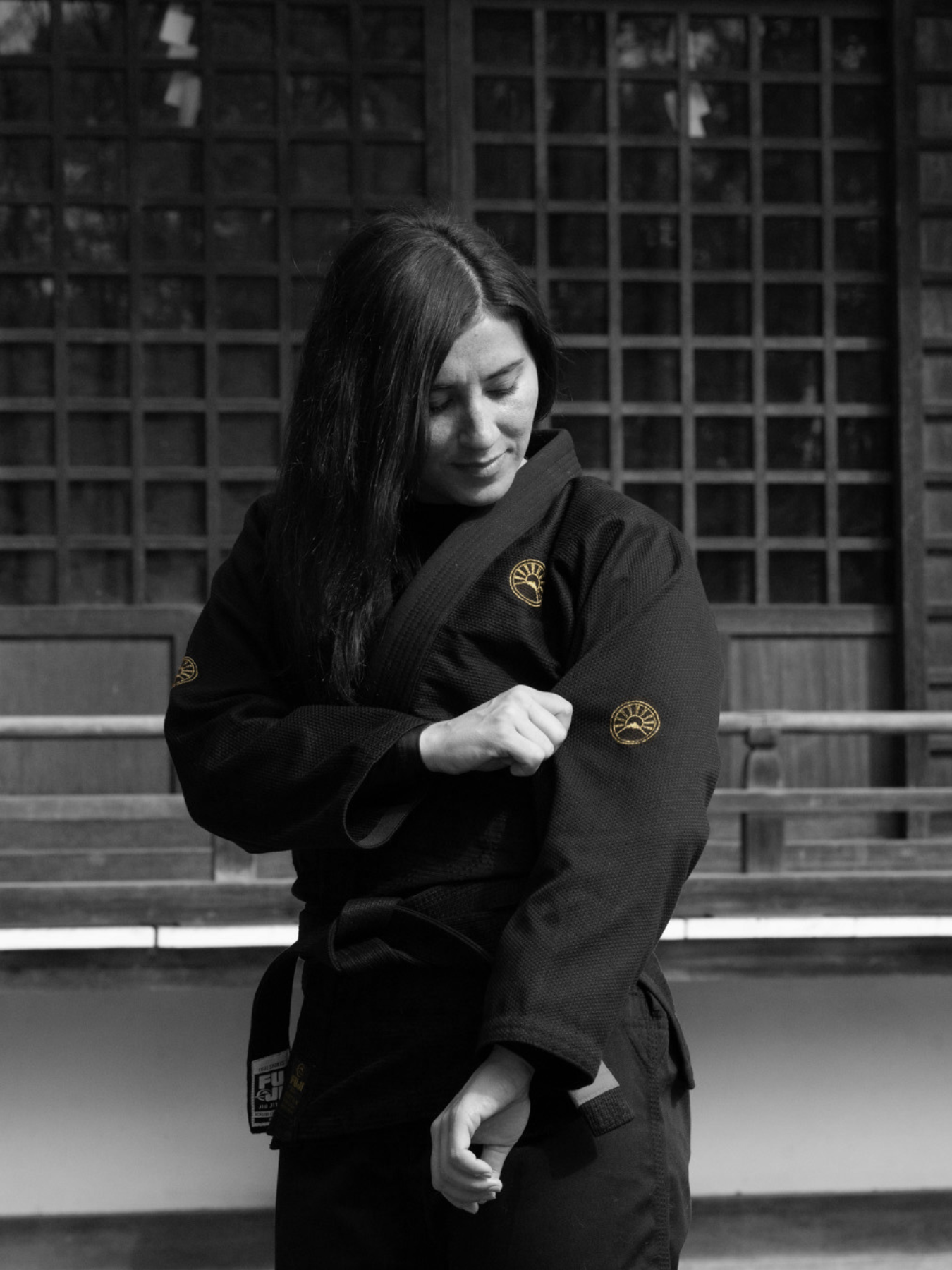 Wearing the Womens Heritage Keisho Lightweight BJJ Gi in black, a woman stands outside adjusting her sleeve, with a wooden building featuring square windows in the background.