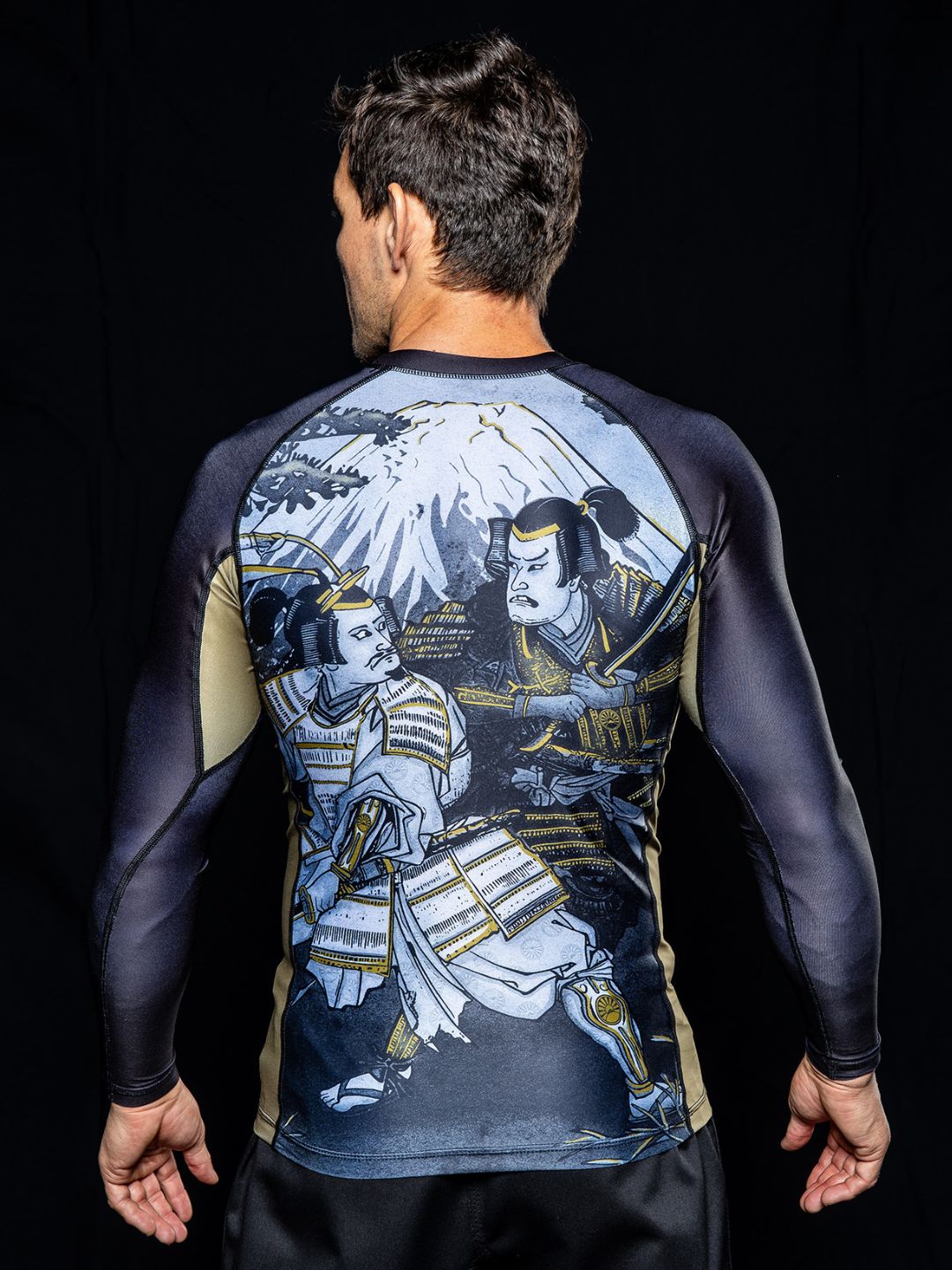 A man stands with his back to the camera, wearing the Heritage Keisho Long Sleeve Rashguard Black, which features a detailed illustration of two samurai and Mount Fuji against a black background.
