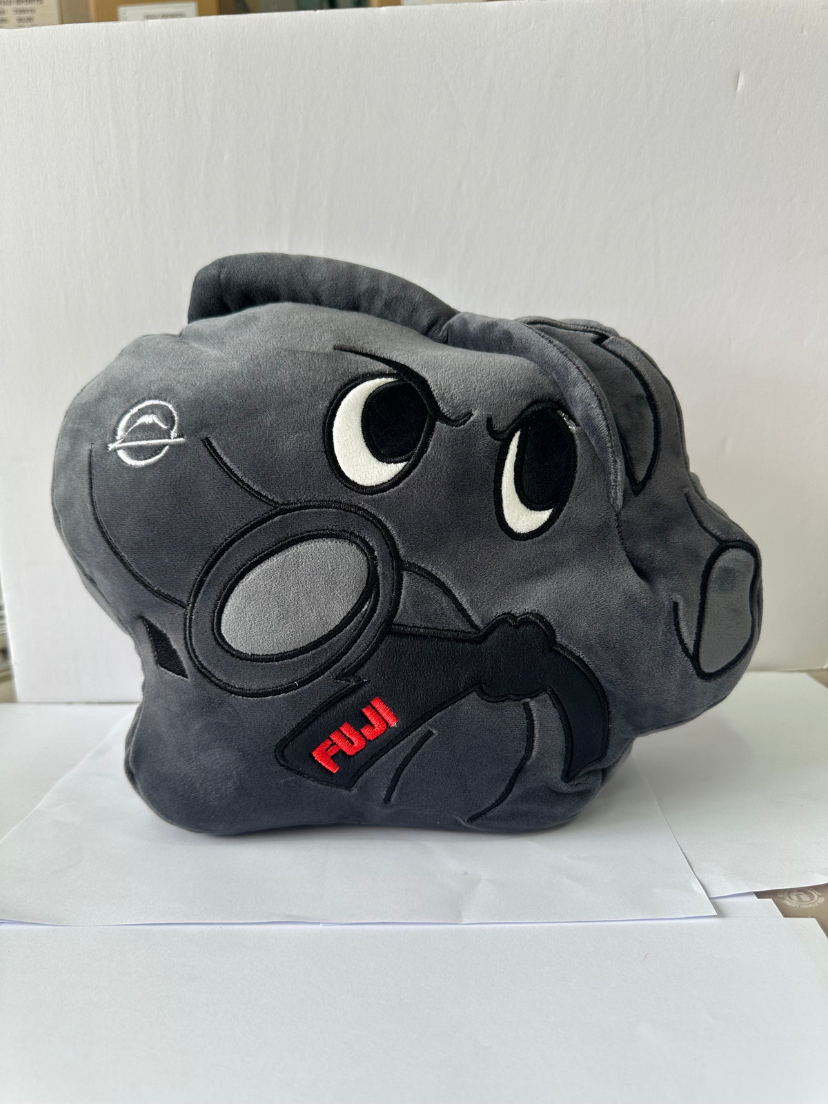The Pochi Piro Pillow Black is a dark gray hippo plush with big black and white eyes, holding a black magnifying glass. “FUJI” is embroidered in red on its body. Set against a plain white background.